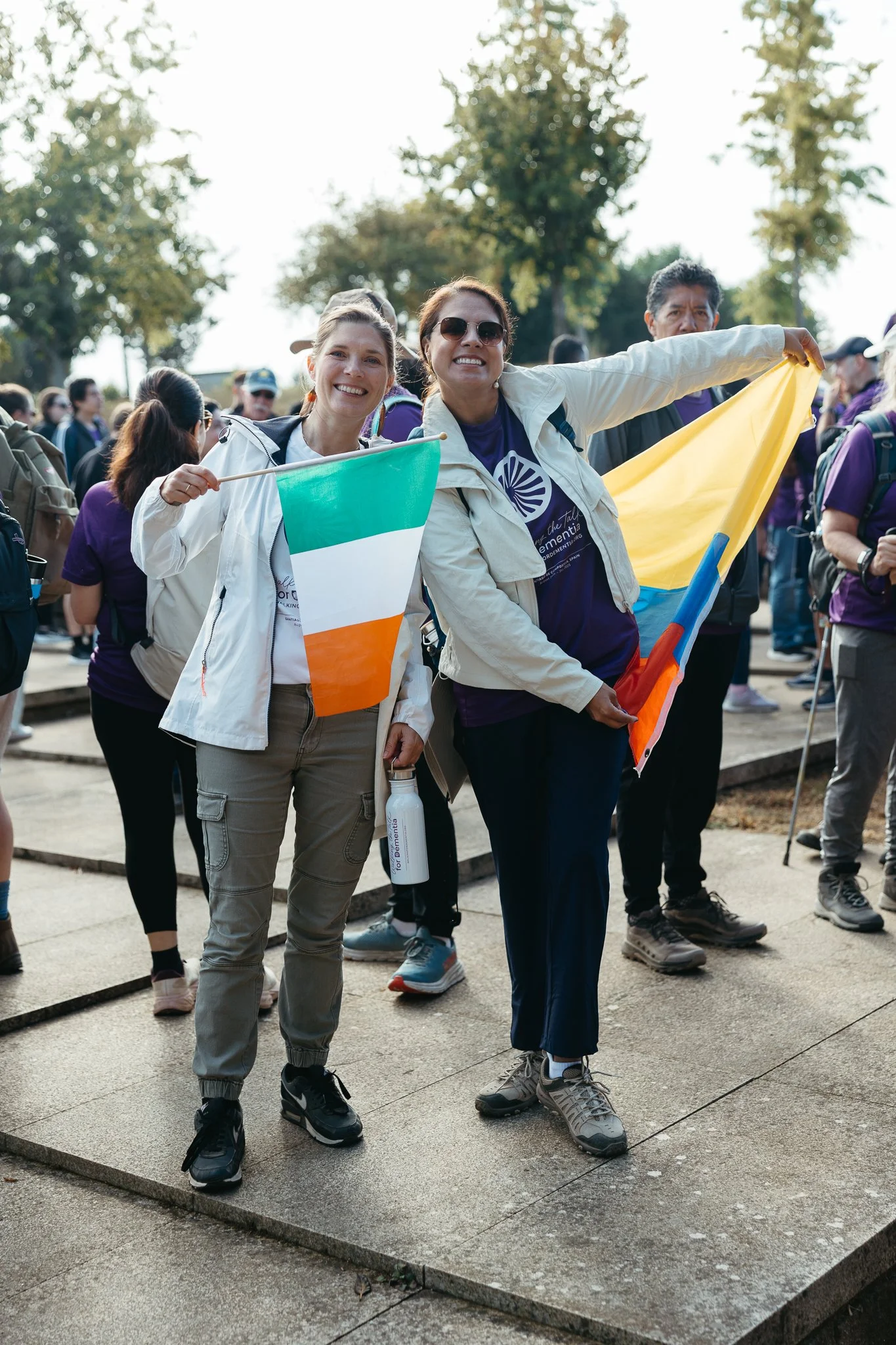 Two women smiling and holding flags, surrounded by a crowd outdoors during daytime.