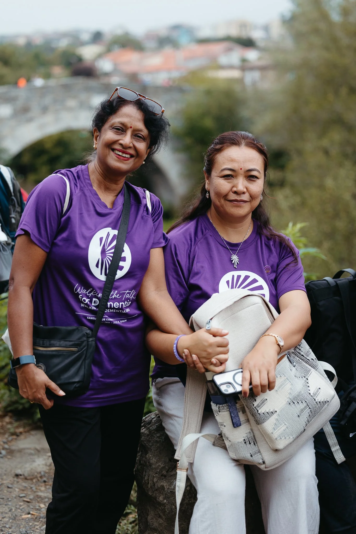 Two women wearing purple shirts smiling outdoors, one with sunglasses on her head, holding hands, with trees and a stone bridge in the background.