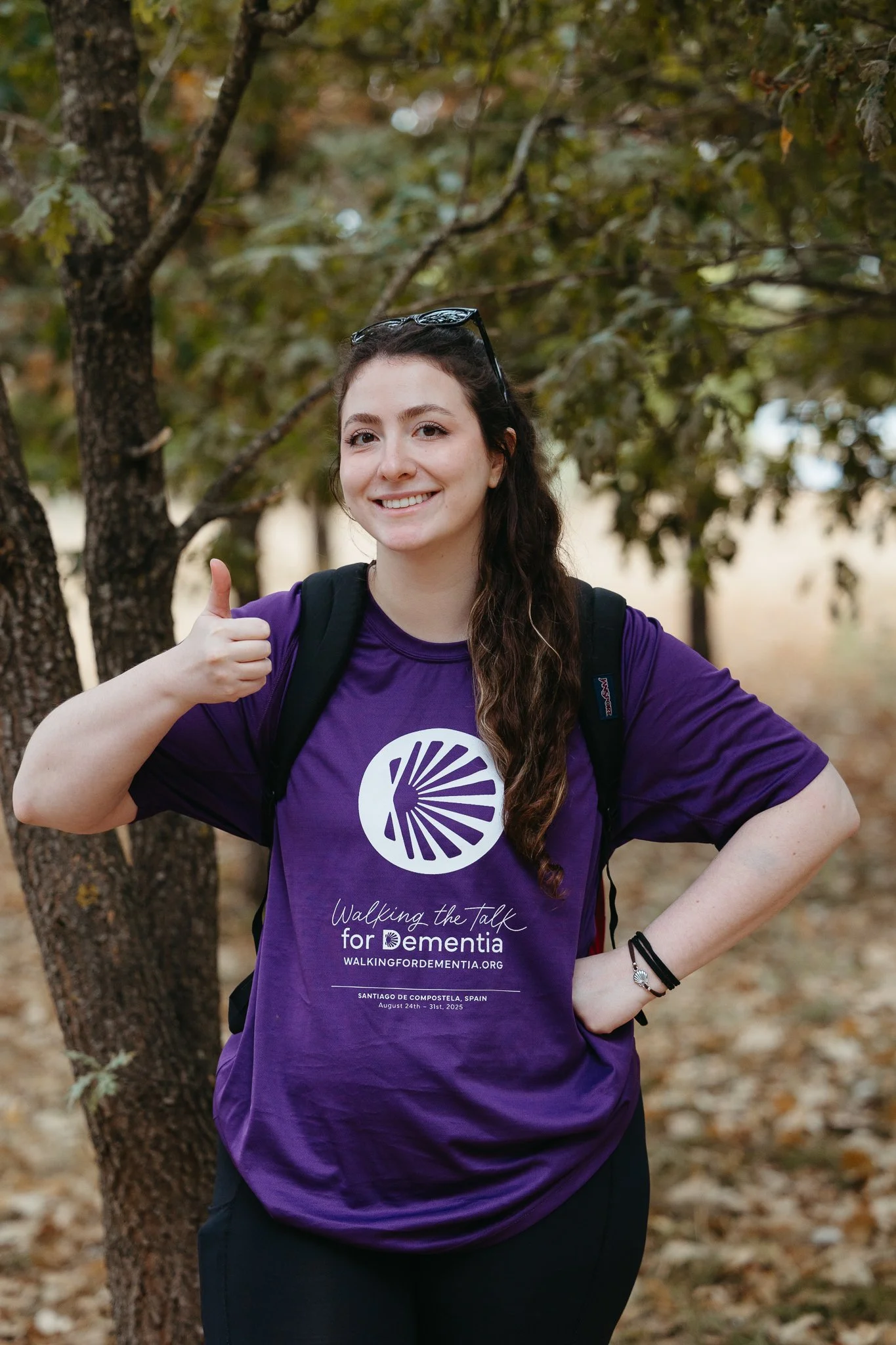 A young woman with long dark hair, wearing a purple t-shirt with event information, giving a thumbs up and smiling outdoors near trees.