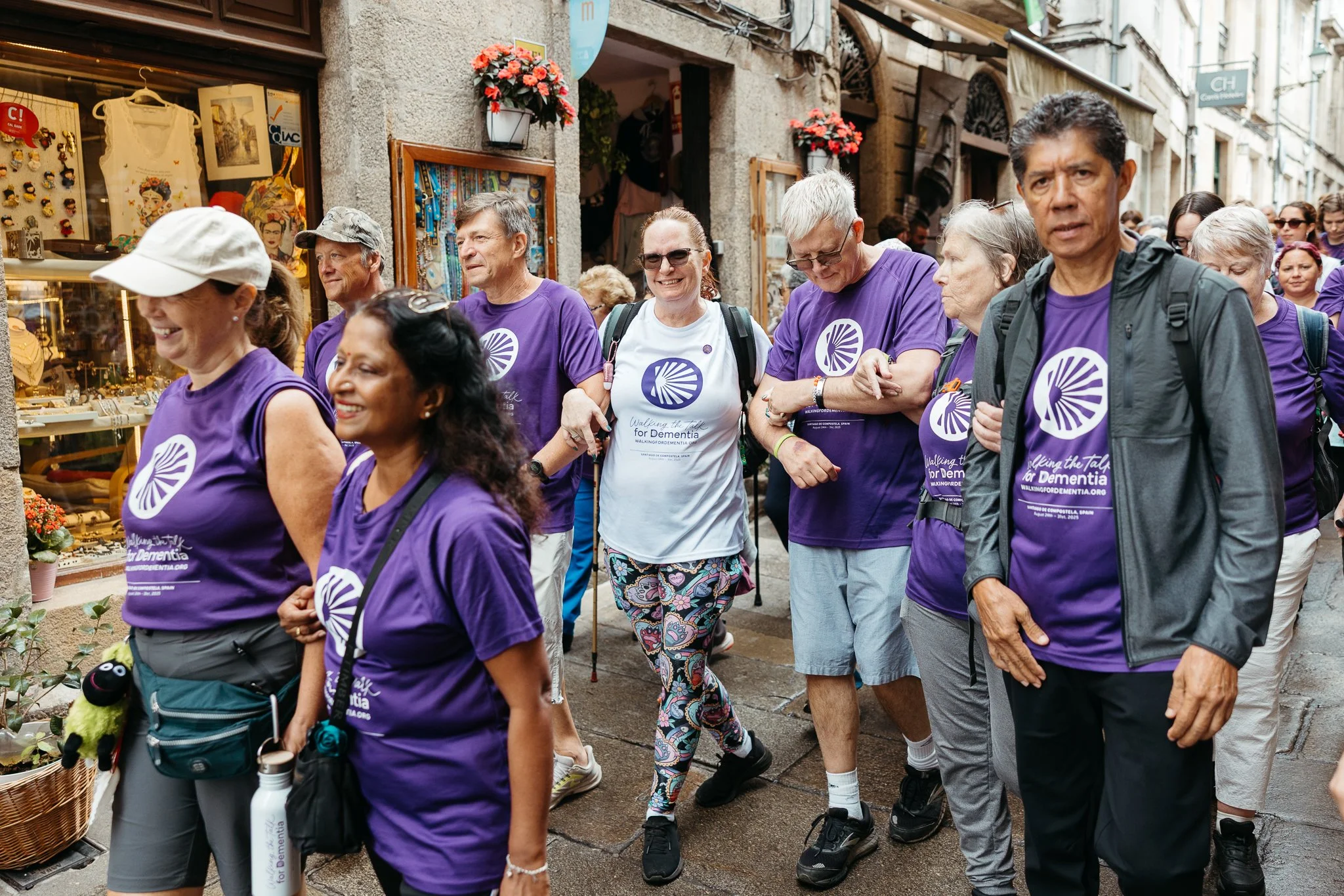 Group of people participating in a walk for dementia awareness in a city street, many wearing purple shirts with a dementia awareness logo, walking past shops decorated with flowers.