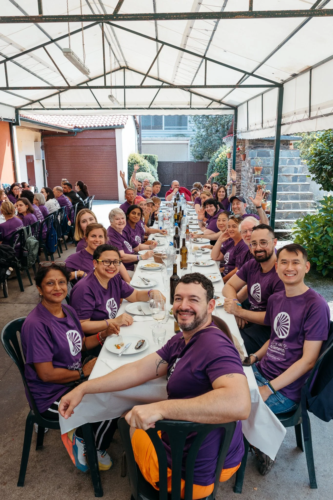 Group of people sitting around a long table under a covered outdoor patio, smiling and waving at the camera, wearing purple shirts, with food and drinks on the table.