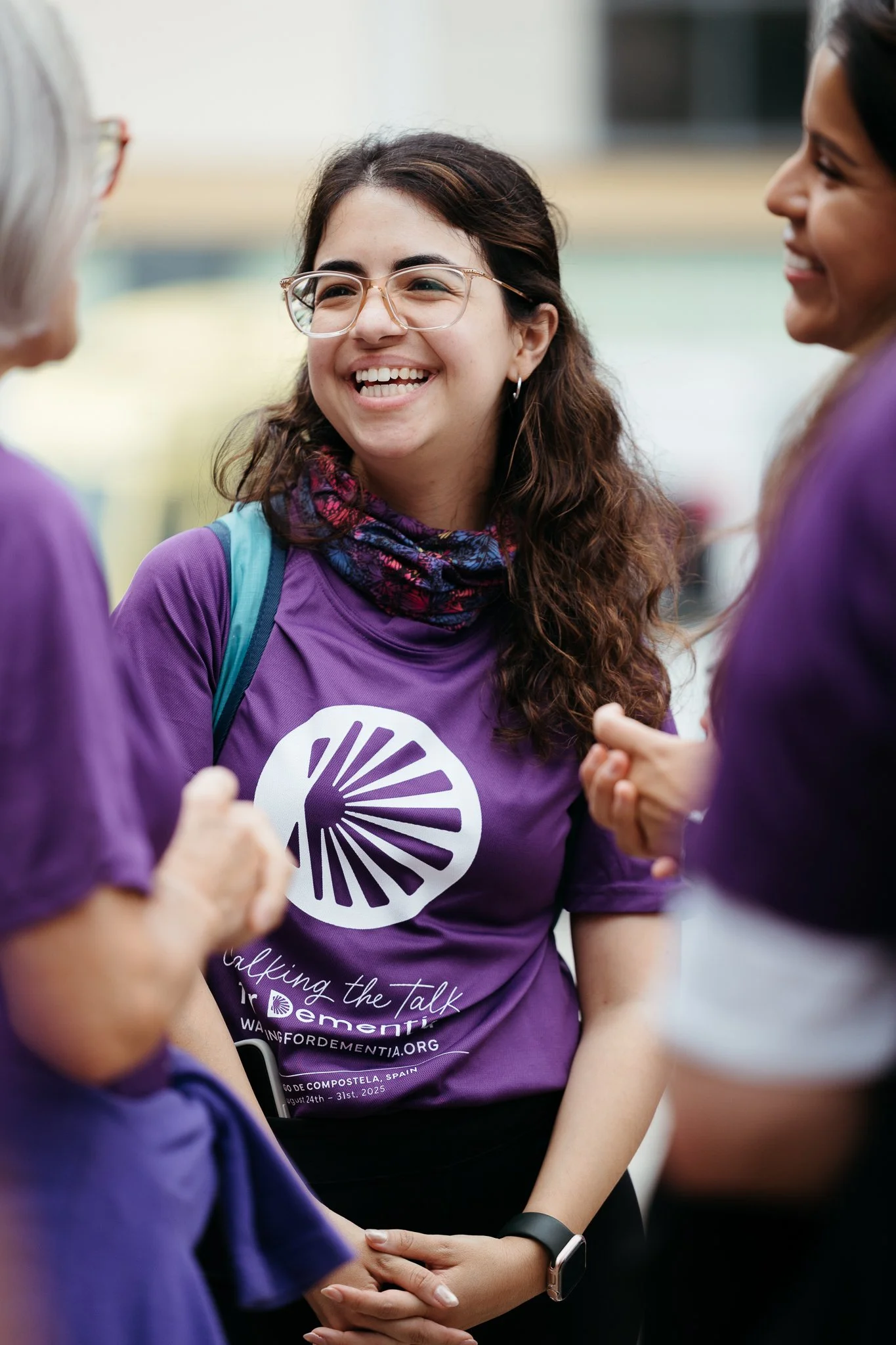A young woman with glasses and brown curly hair smiling and talking with two women, all wearing purple shirts with a logo and text supporting dementia awareness.