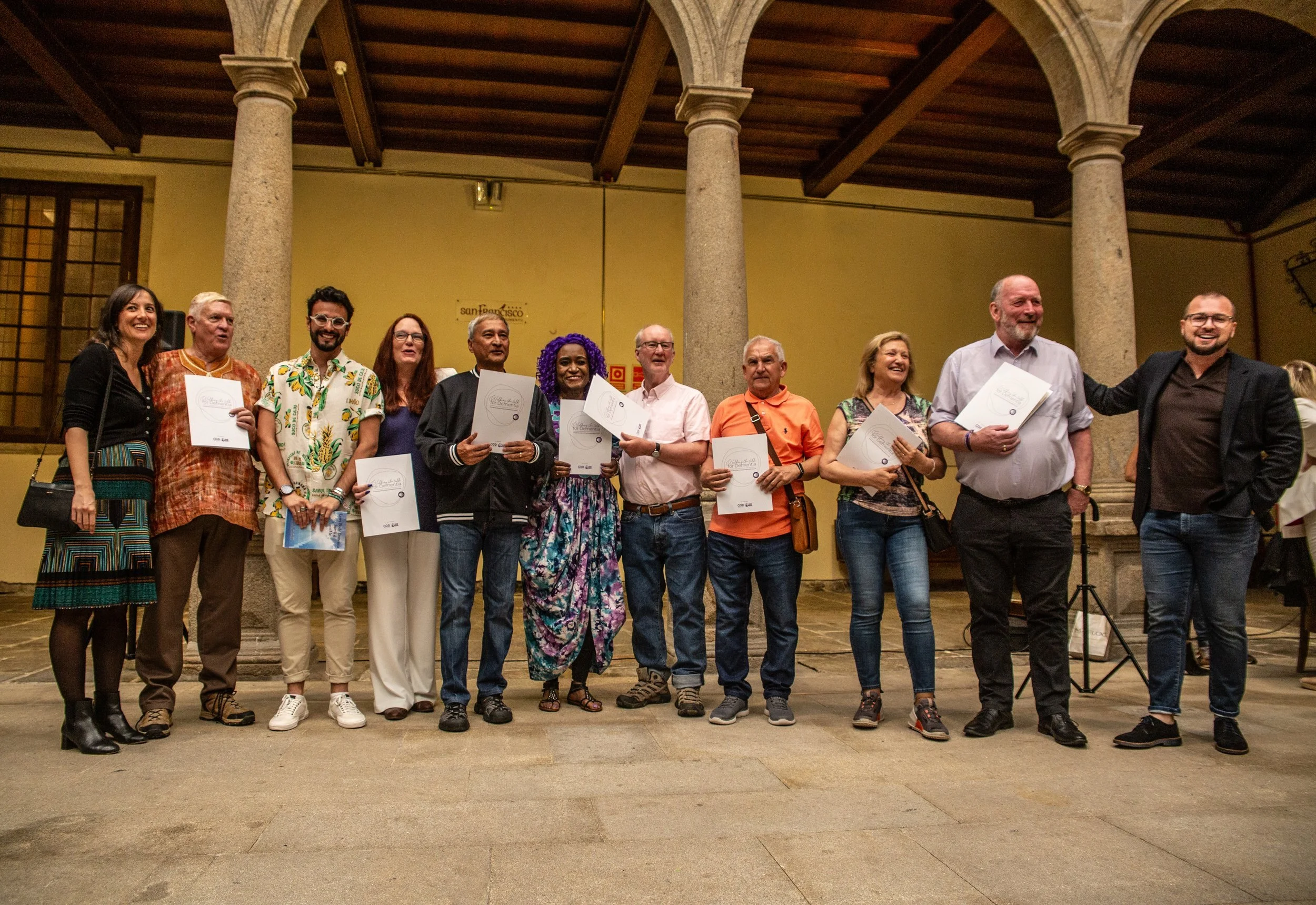 Group of people standing on stage holding certificates, celebrating at an indoor event with arches and columns in the background.