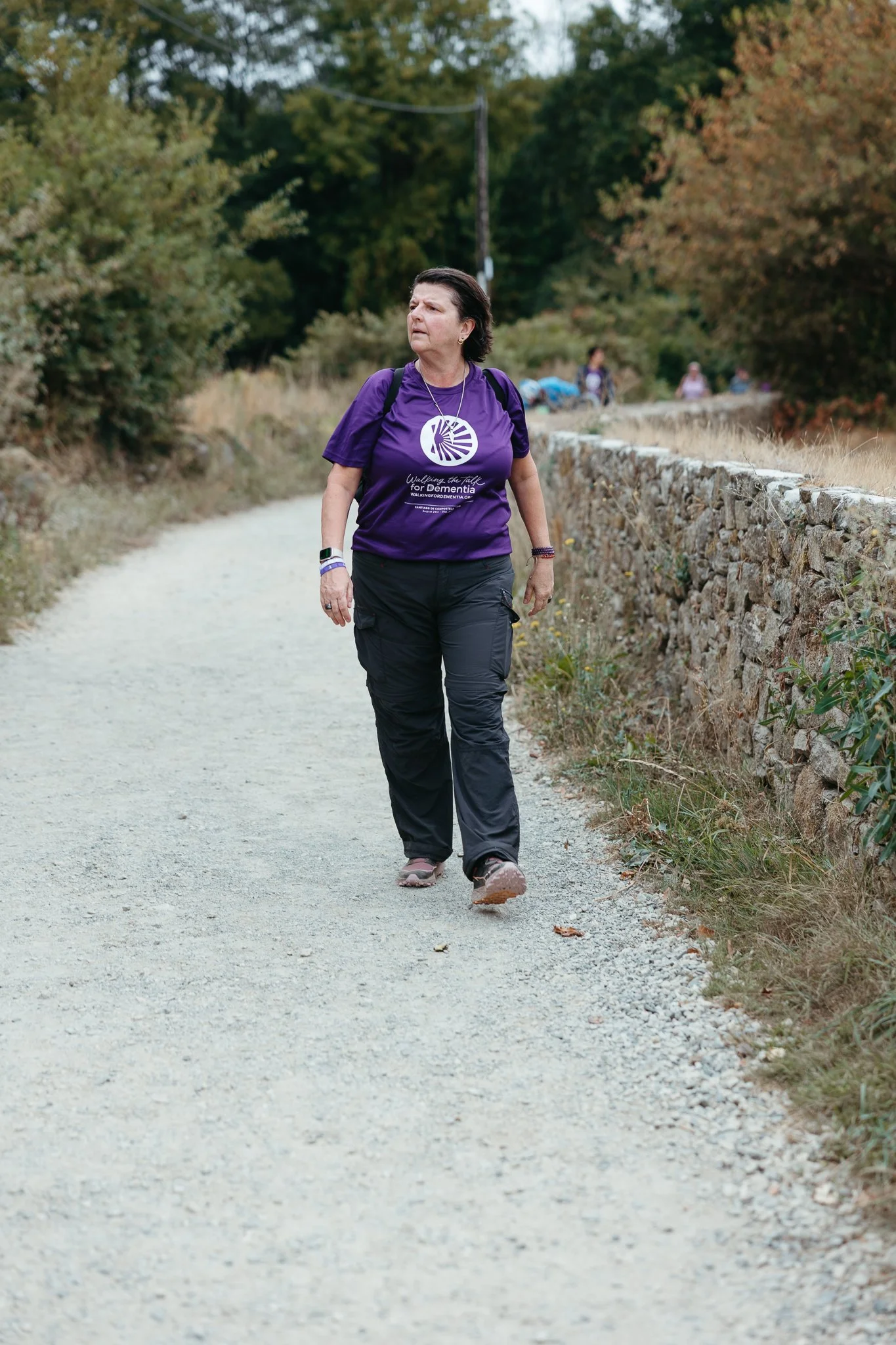 A woman walking on a gravel path in a park, wearing a purple T-shirt and black pants.