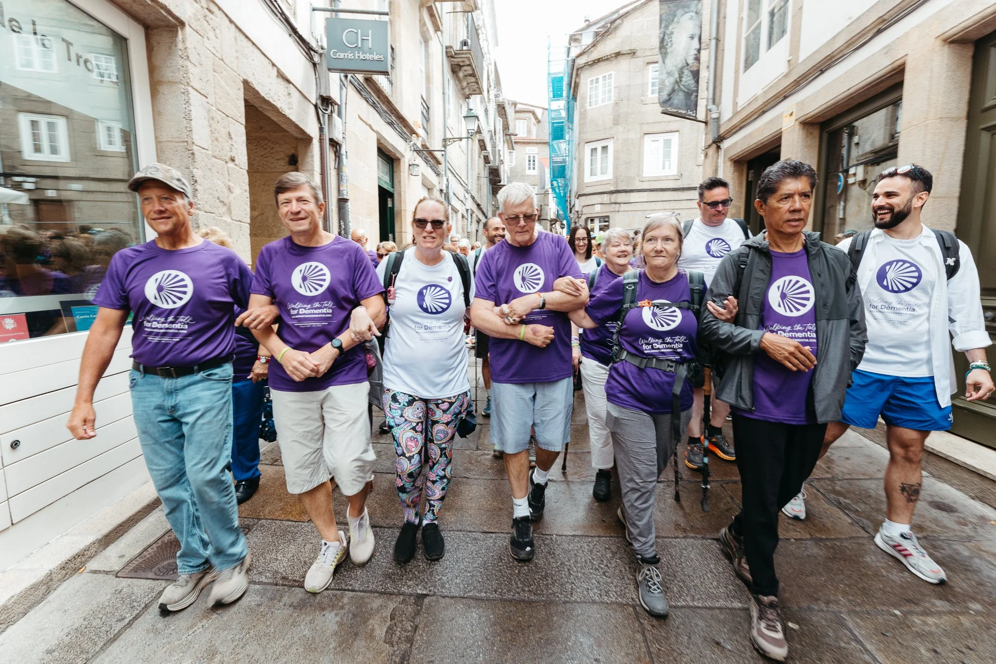 A group of people walking together on a city street, many wearing purple or white shirts with a logo and the text 'Walking the Talk for Dementia'.