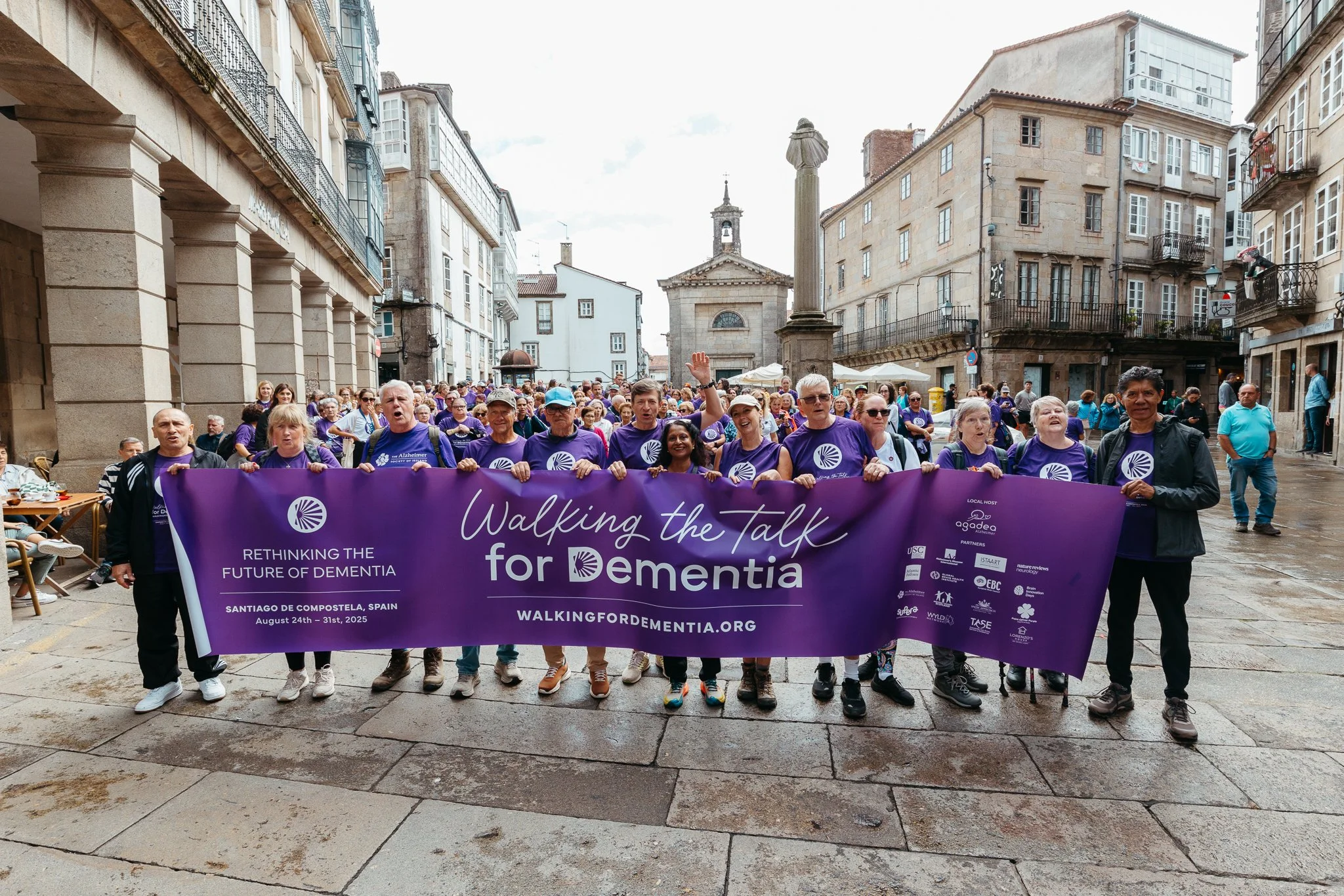 Group of people holding a purple banner during a walk event in Santiago de Compostela, Spain, advocating for dementia awareness, with historic buildings in the background.
