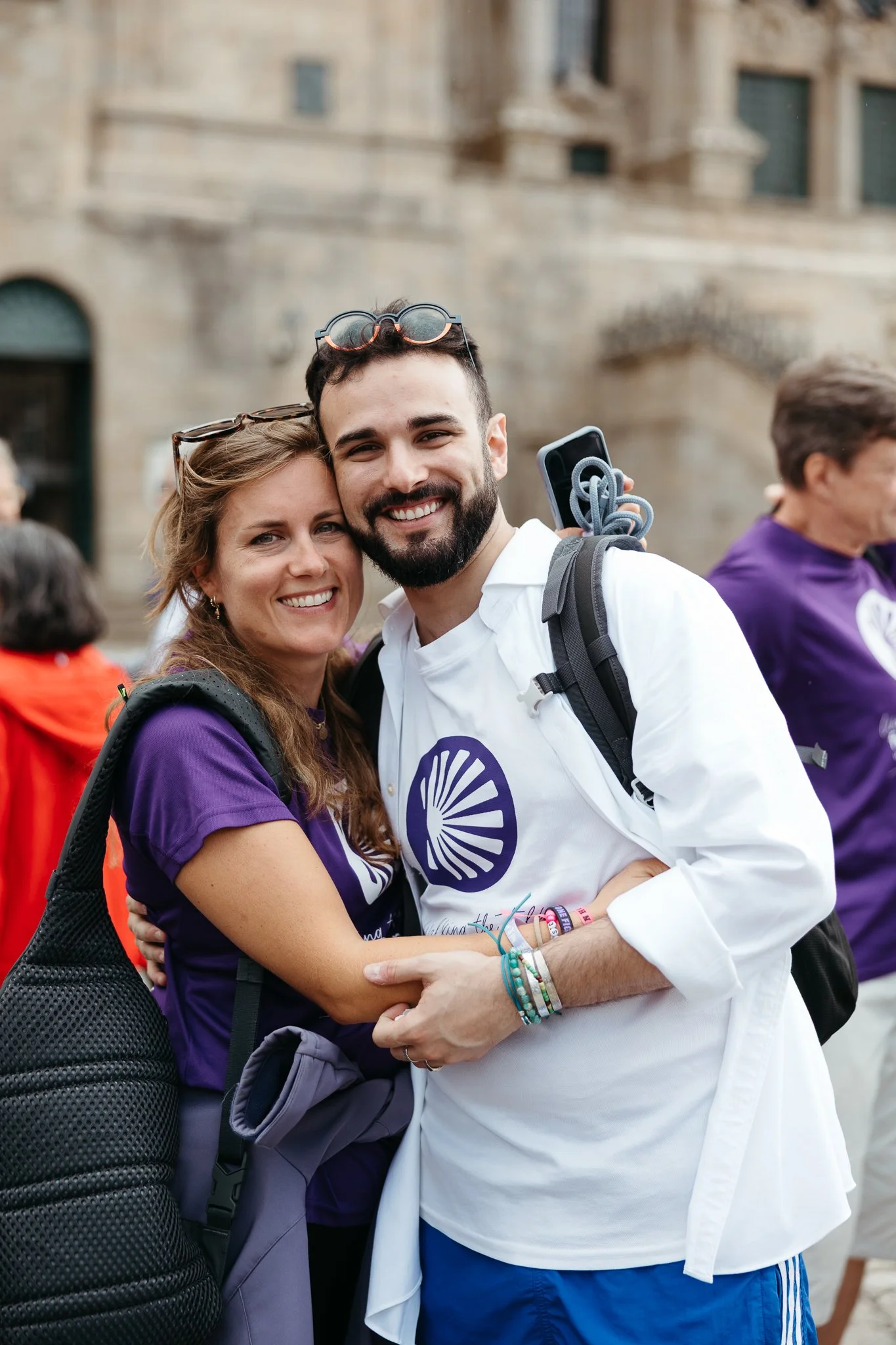 A smiling young couple hugging each other, wearing purple shirts with a white logo, standing outdoors in front of a historic building, at a public event or gathering.