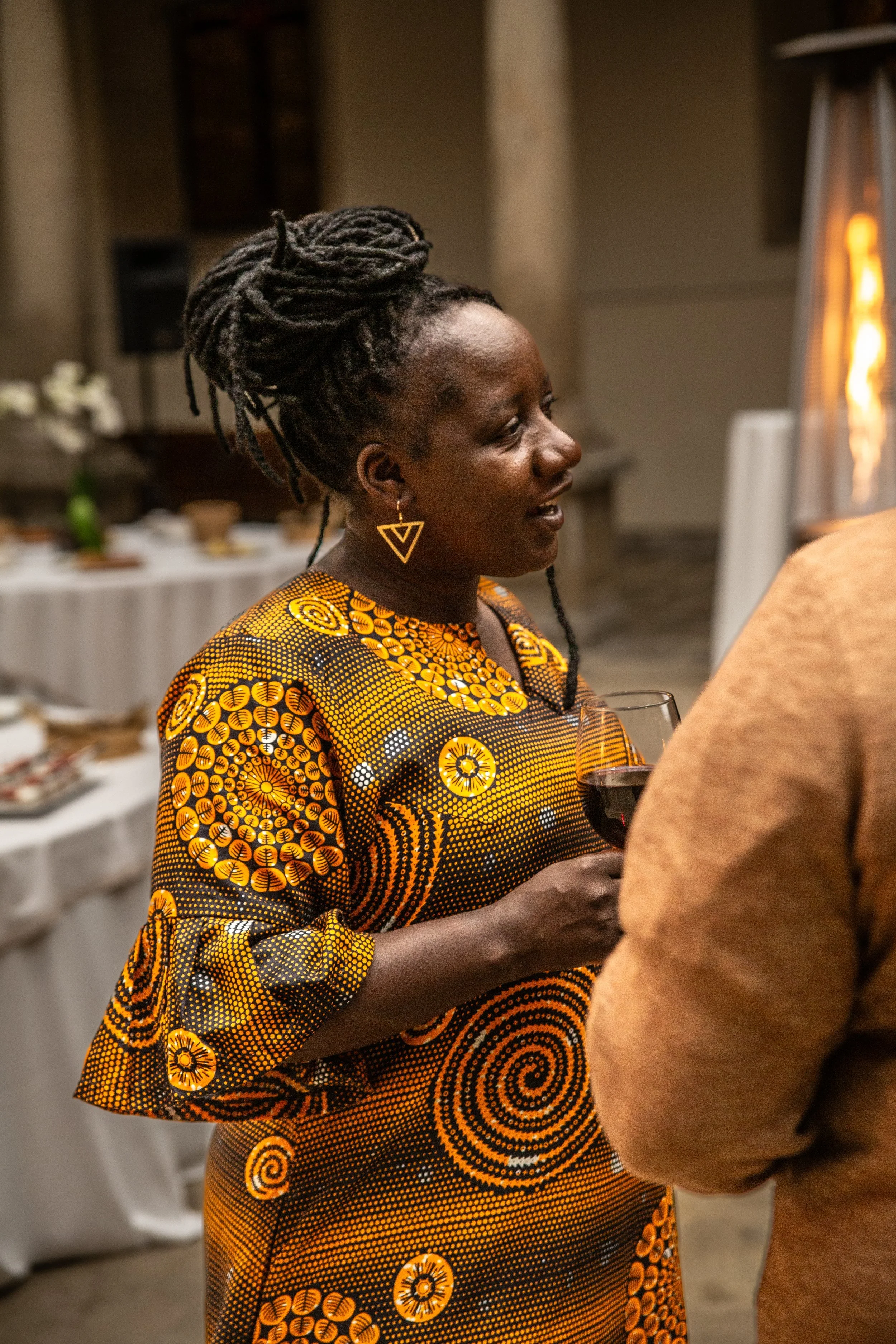 A woman with dark skin and dreadlocks wearing a colorful, patterned dress, holding a glass of red wine at a social gathering.