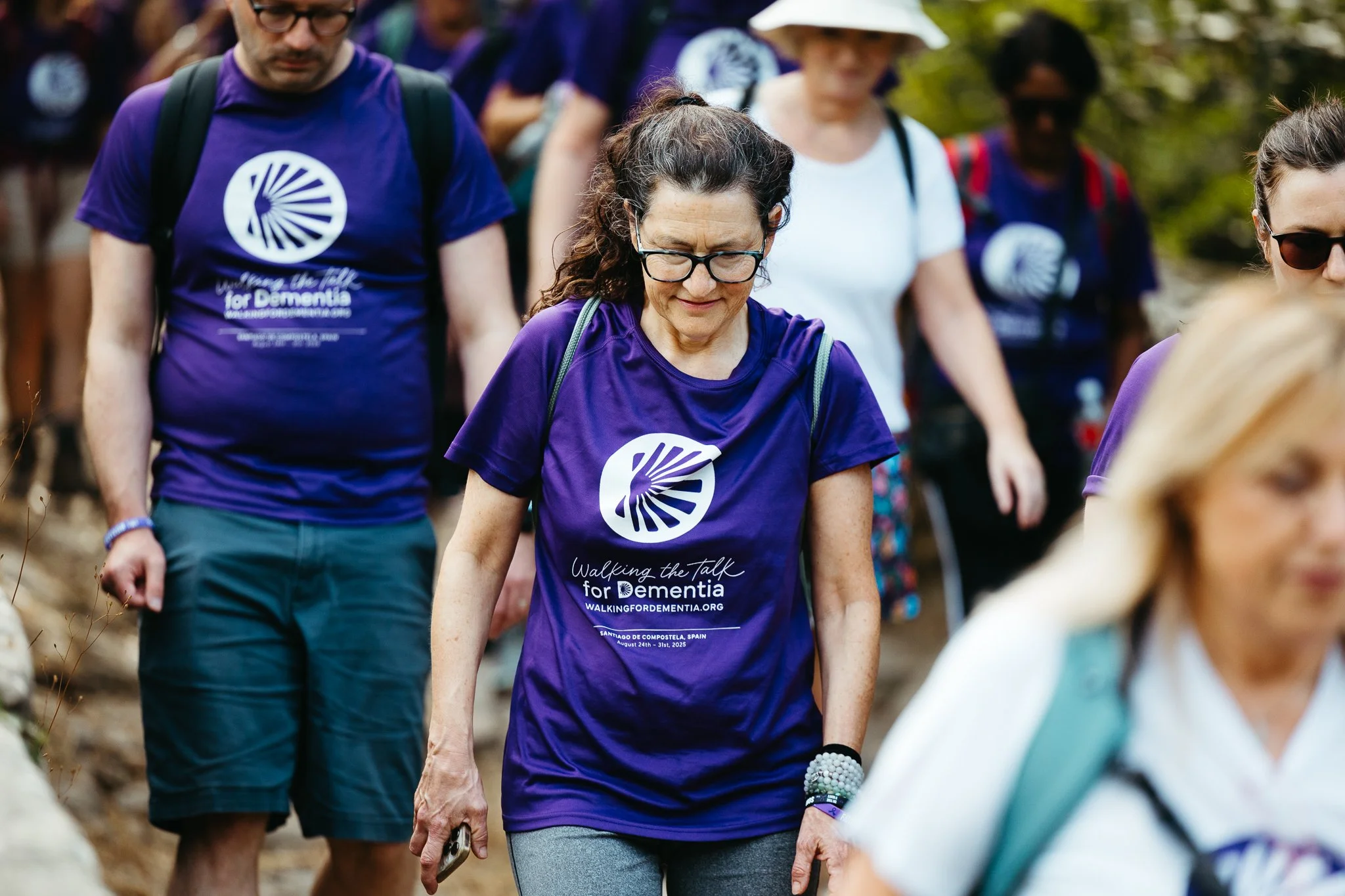 A group of people walking outdoors, wearing purple t-shirts with a logo and text promoting a dementia awareness walk, with some wearing backpacks and sunglasses.