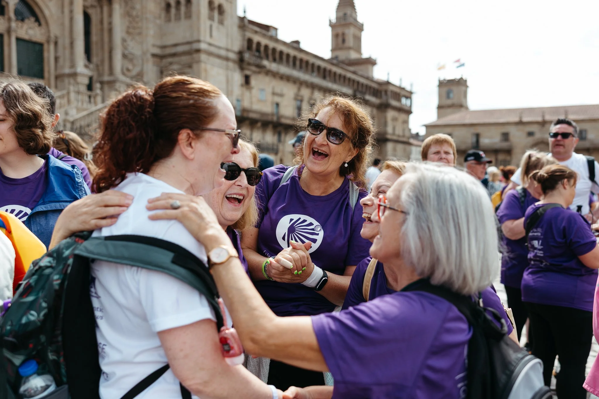 Group of women and men smiling and laughing, gathered outdoors in front of historic buildings, some wearing purple shirts with a logo, engaging in conversation and group hug.
