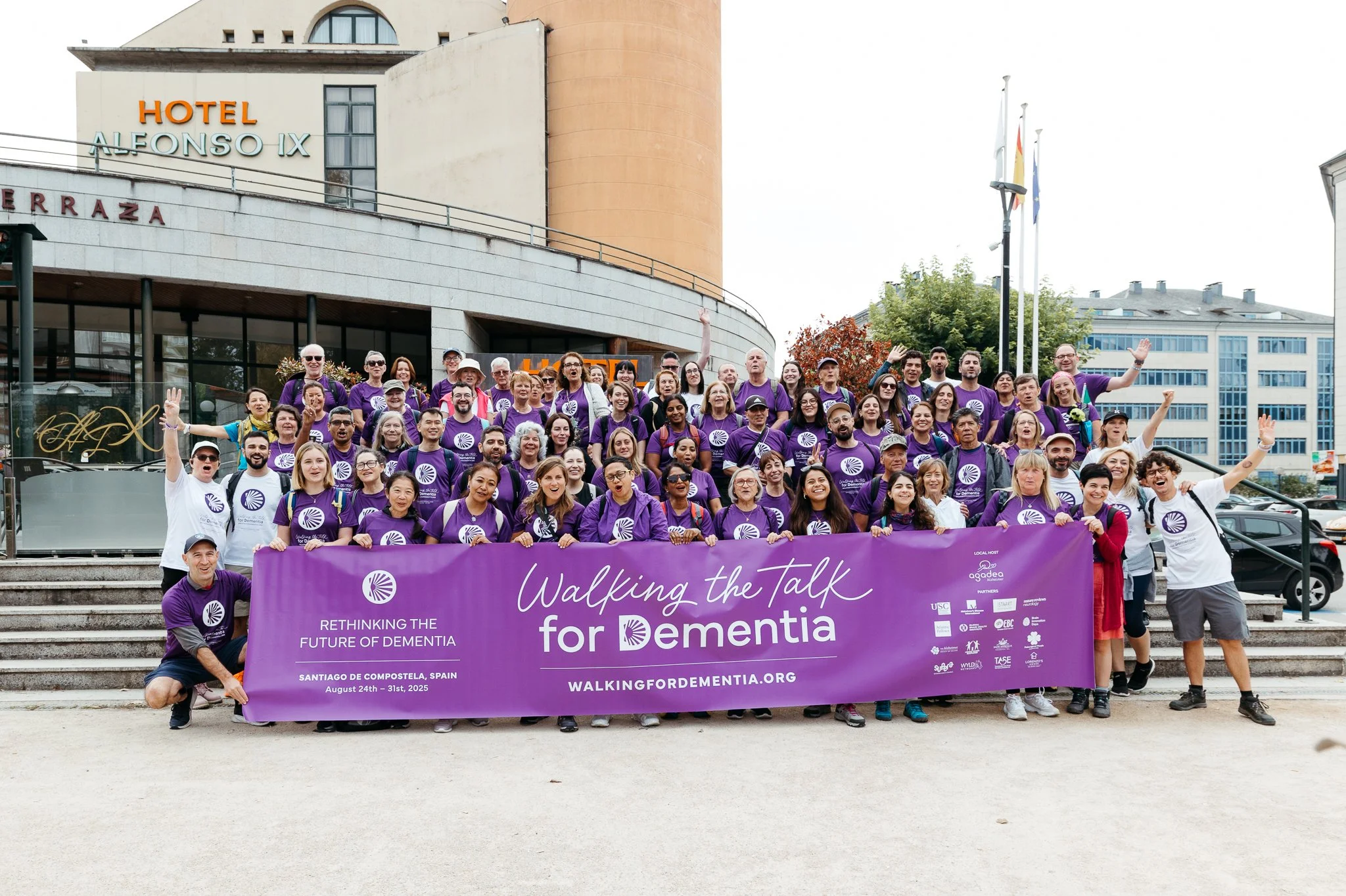 Group of people in purple shirts holding a large purple banner outside a building with hotel signs, participating in a dementia awareness walk in Santiago de Compostela, Spain.