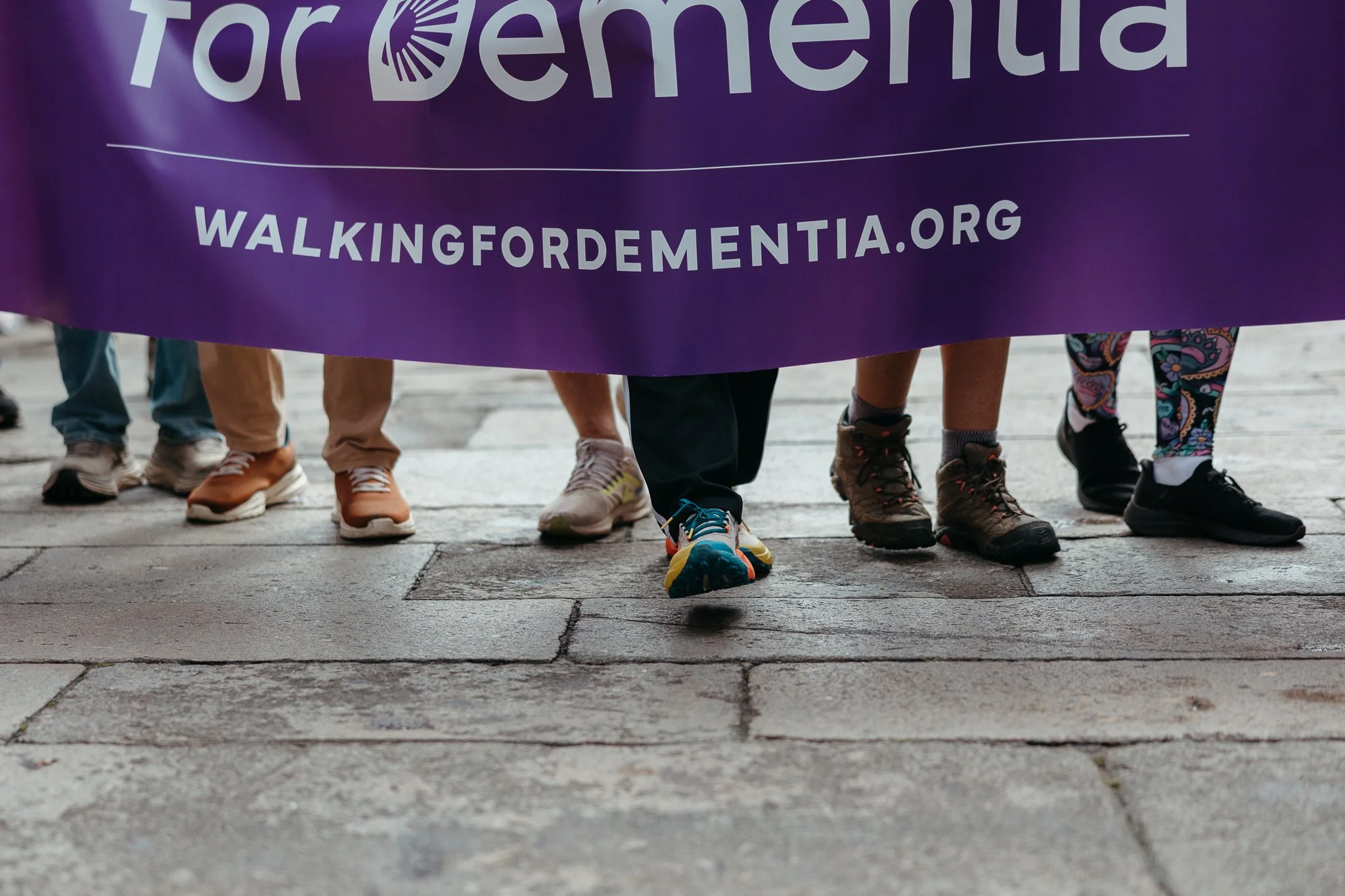 People holding a purple banner with the words 'for DEMENTIA' and the website 'WALKINGFORDEMENTIA.ORG,' standing on a stone sidewalk.