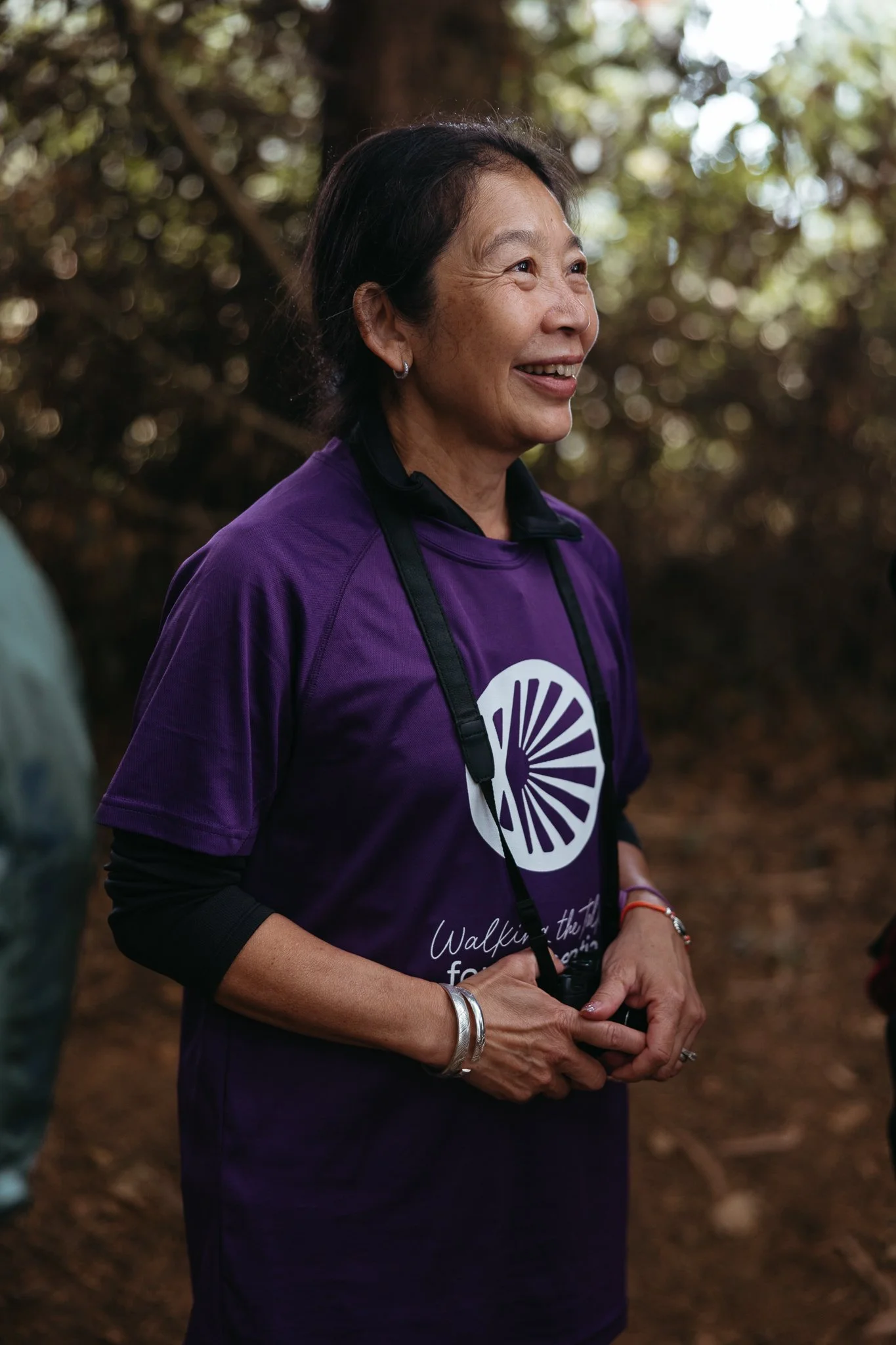 Smiling woman wearing a purple t-shirt with a camera hanging around her neck, standing outdoors with trees in the background.