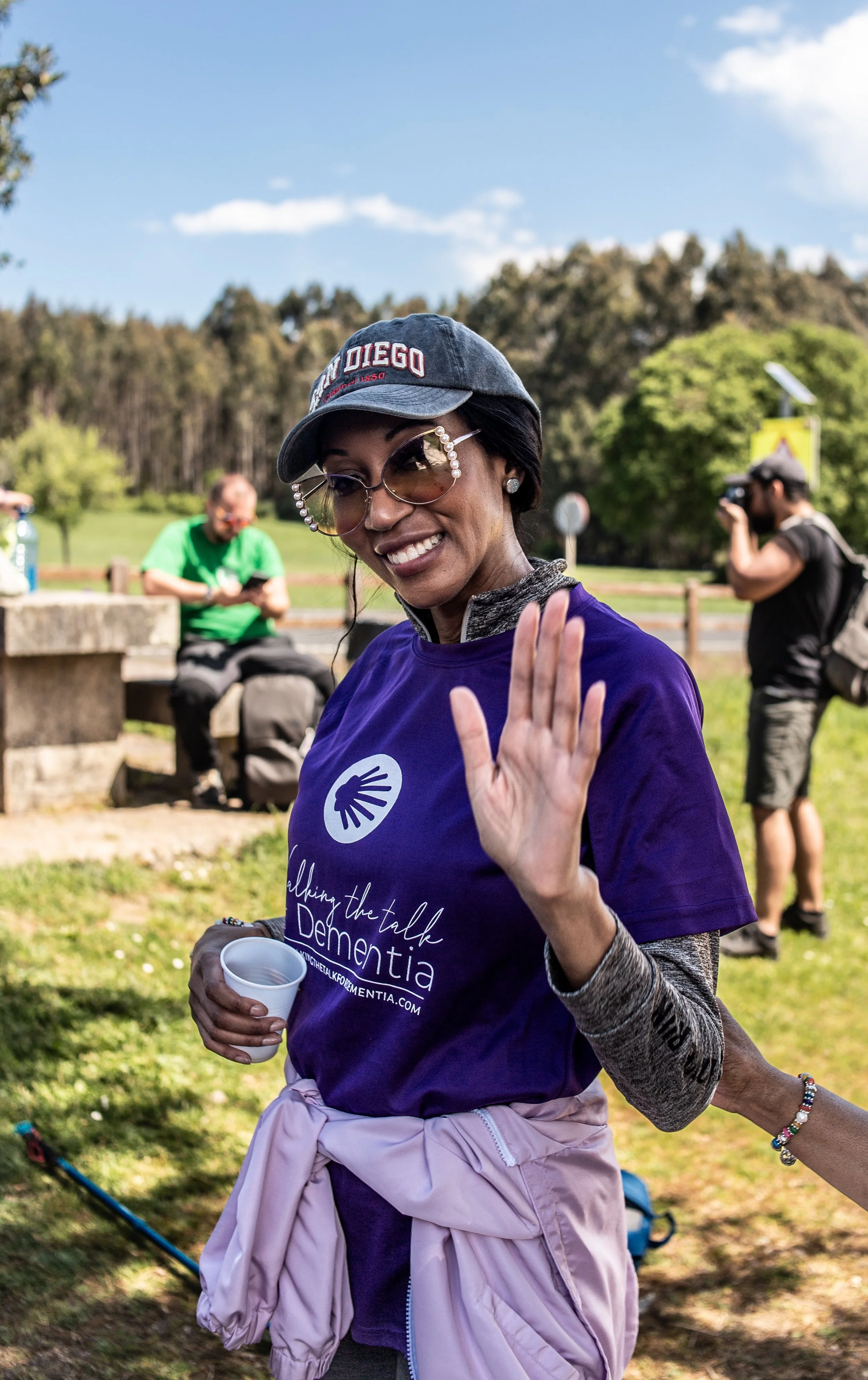 Smiling woman wearing sunglasses, a purple t-shirt with 'Walking the Talk of Dementia' written on it, a gray cap with 'San Diego' embroidered on it, holding a cup and waving outdoors on a sunny day.
