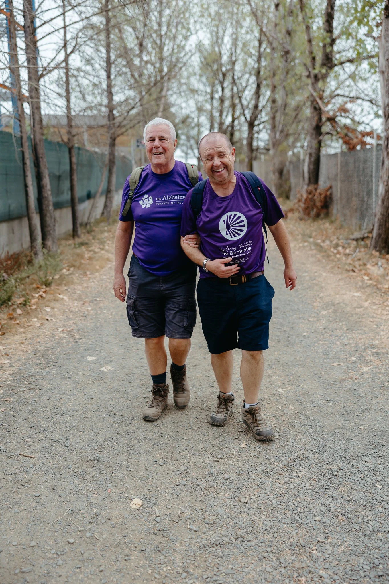 Two men walking outdoors on a dirt path, smiling, wearing purple T-shirts and shorts, with backpacks, surrounded by trees.