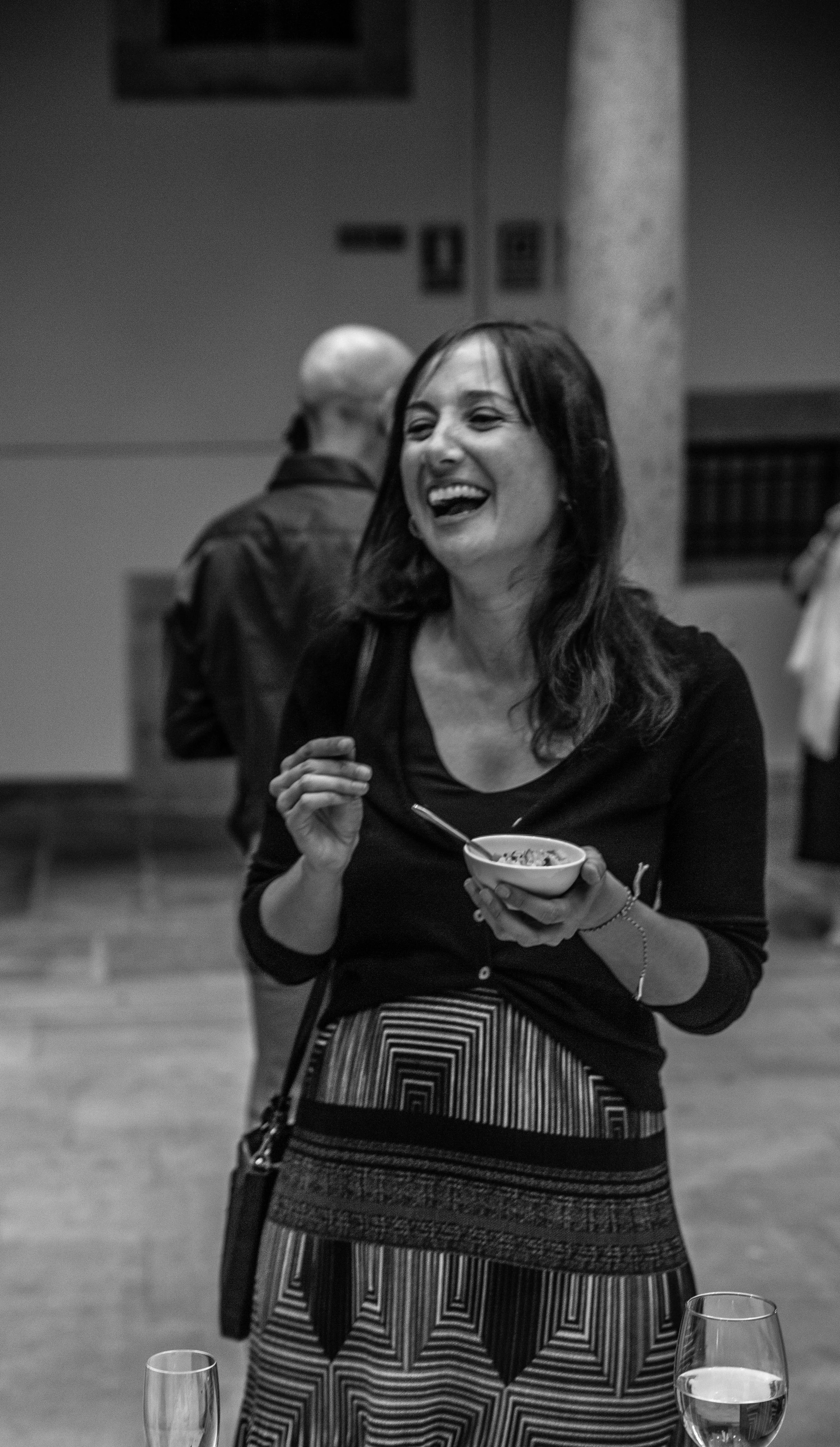 A woman with dark hair wearing a black top and a patterned skirt, smiling and holding a bowl and spoon, at an indoor social gathering.