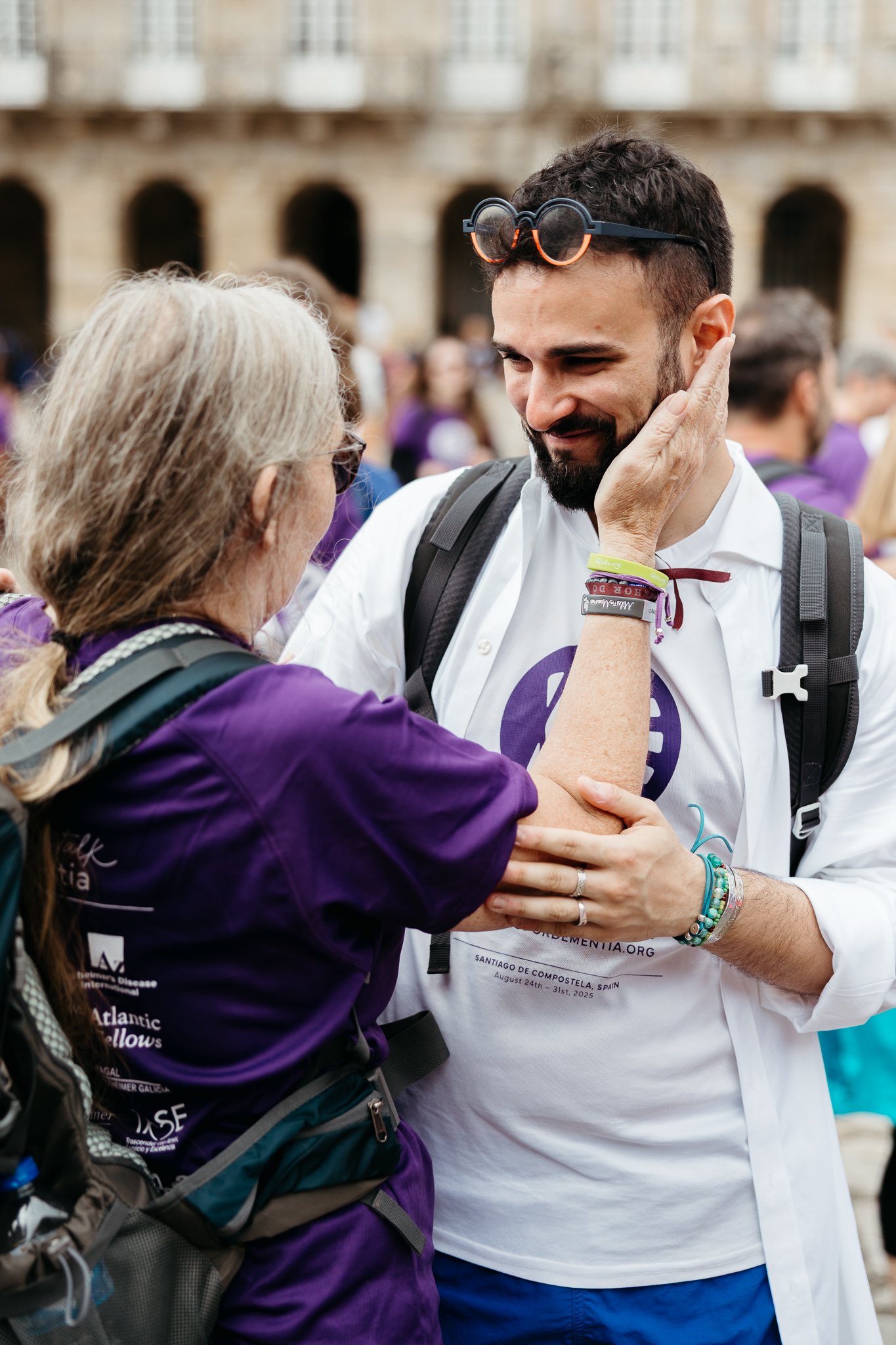 A man with dark hair and a beard, wearing a white shirt and backpacks, lovingly touches the face of an older woman with long white hair, who is wearing a purple shirt, during an outdoor event.