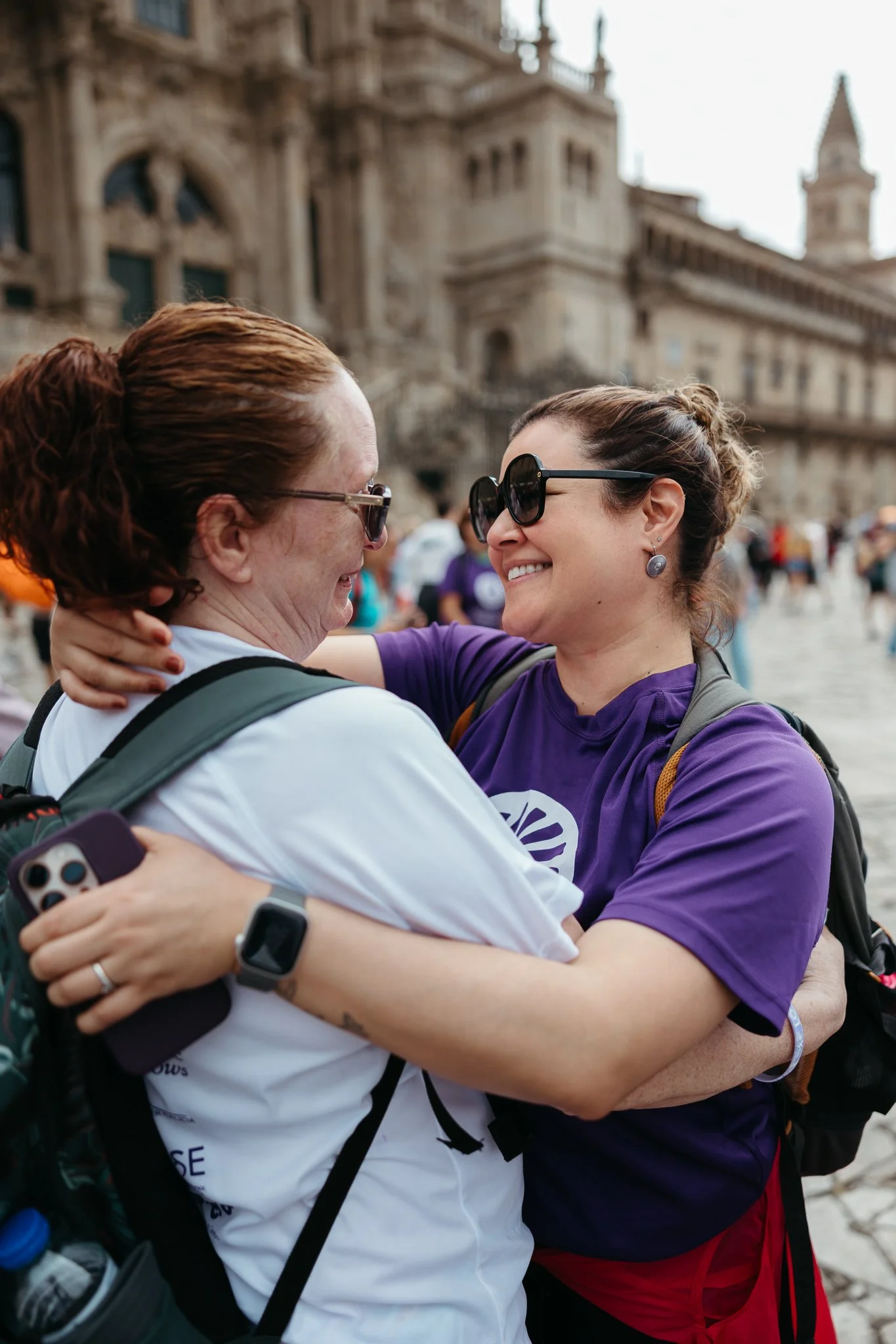 Two women hugging and smiling, wearing sunglasses, in front of a historic building with ornate architecture, with other people in the background.