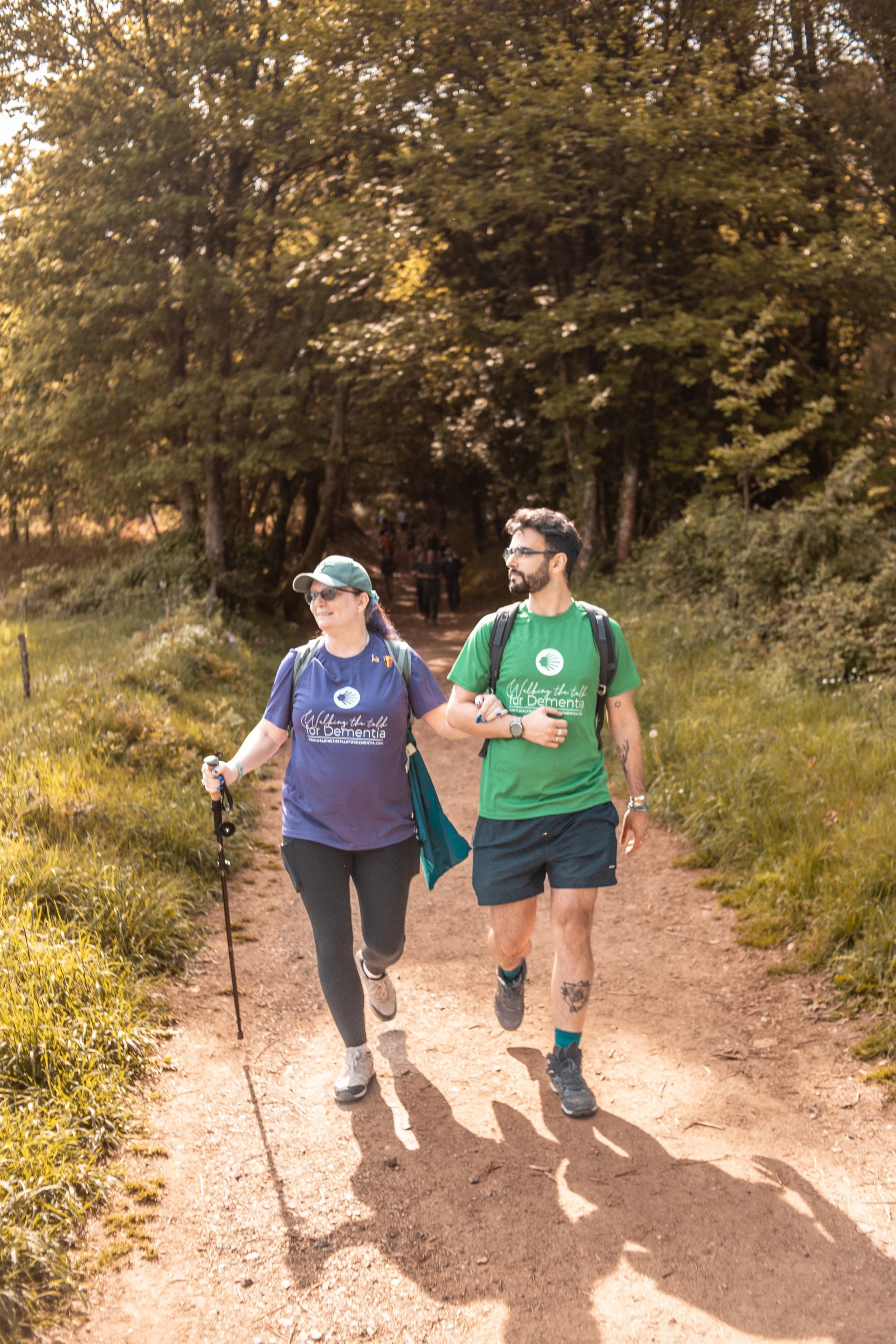 Two people walking on a dirt trail in a wooded area during daylight. The woman on the left is smiling, wearing a blue T-shirt, black leggings, a cap, and sunglasses, and carrying a walking pole. The man on the right is wearing a green T-shirt and bla