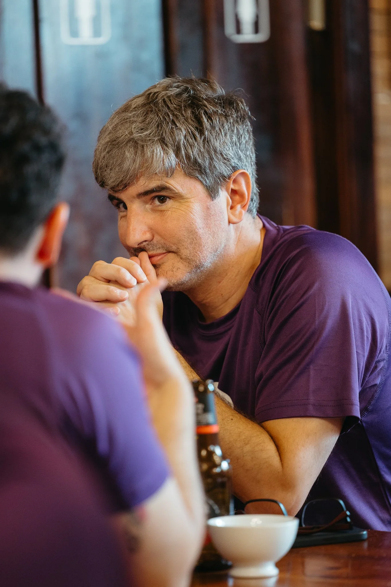 A man with gray hair and a beard leaning on a table, looking thoughtfully at another person with blurred dark hair and purple shirt, in a casual indoor setting.