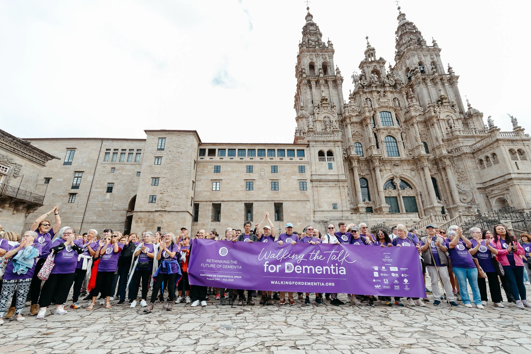 A large group of people participating in a walk for dementia awareness, holding a purple banner that reads 'Walking the Talk for Dementia' in front of a historic cathedral with detailed stone architecture in Santiago de Compostela, Spain.