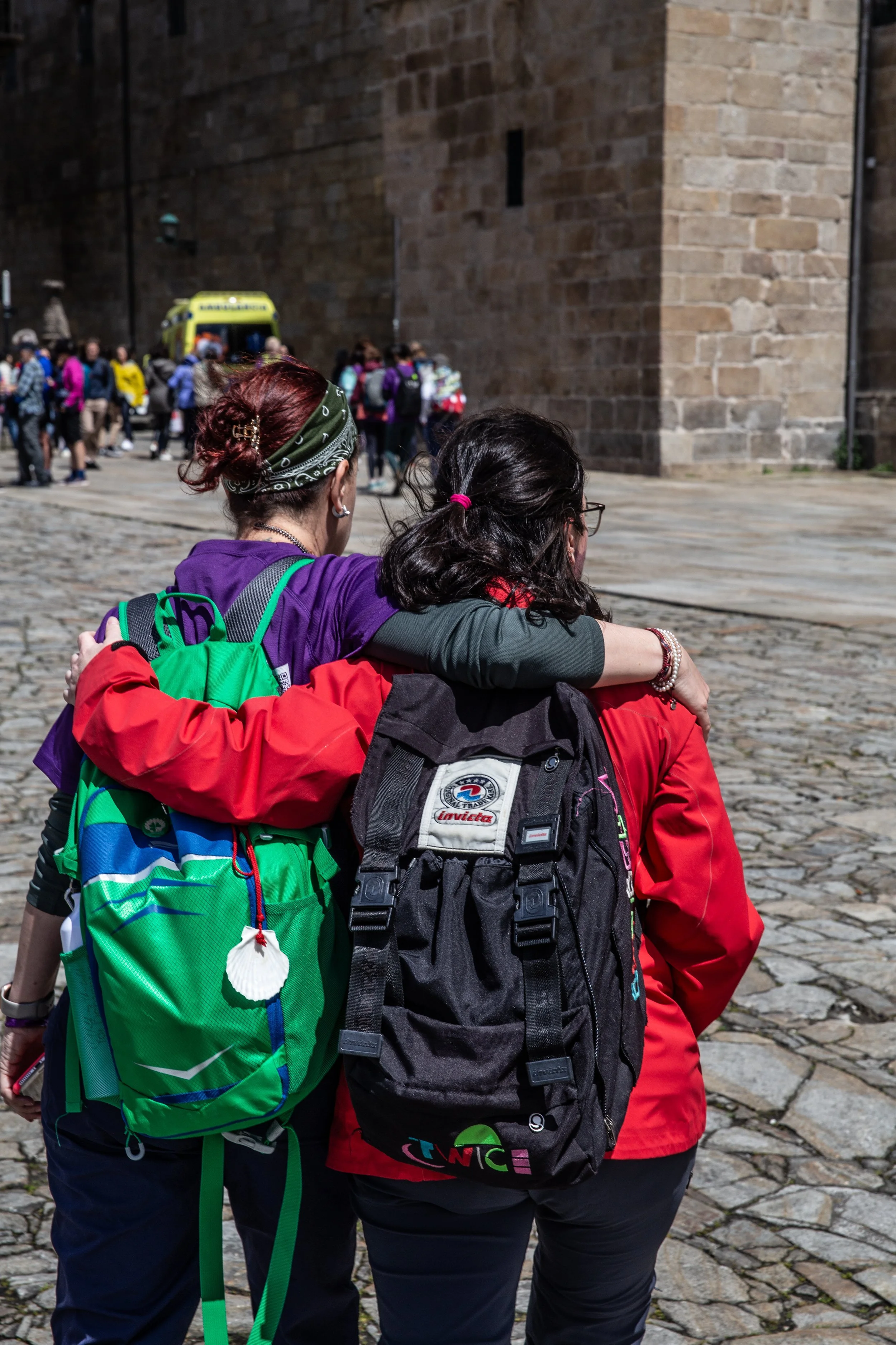 Two women with backpacks hugging each other in a cobblestone street, with a crowd and an ambulance in the background.