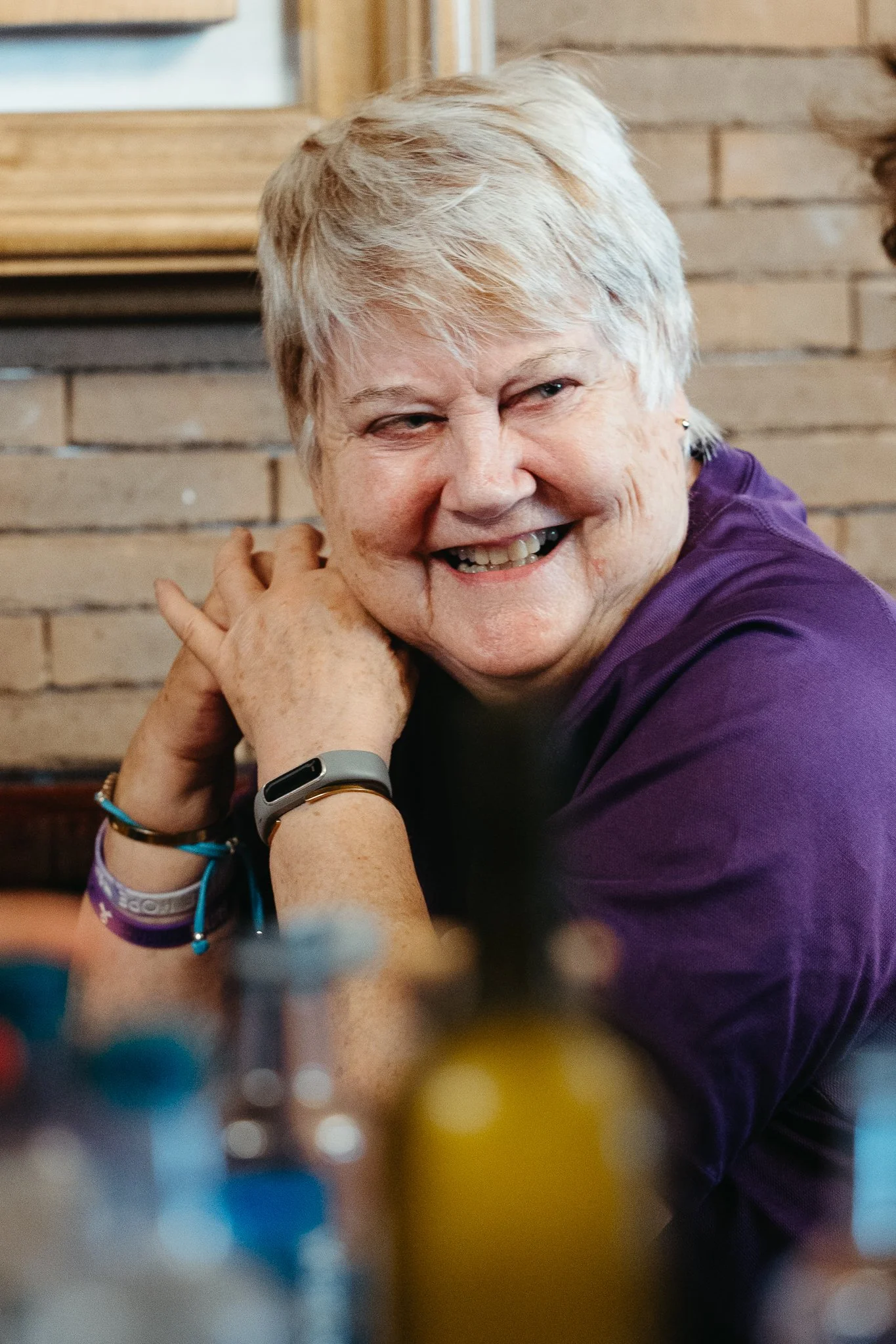 An elderly woman with short gray hair, smiling and resting her head on her hands, sitting in front of a brick wall.