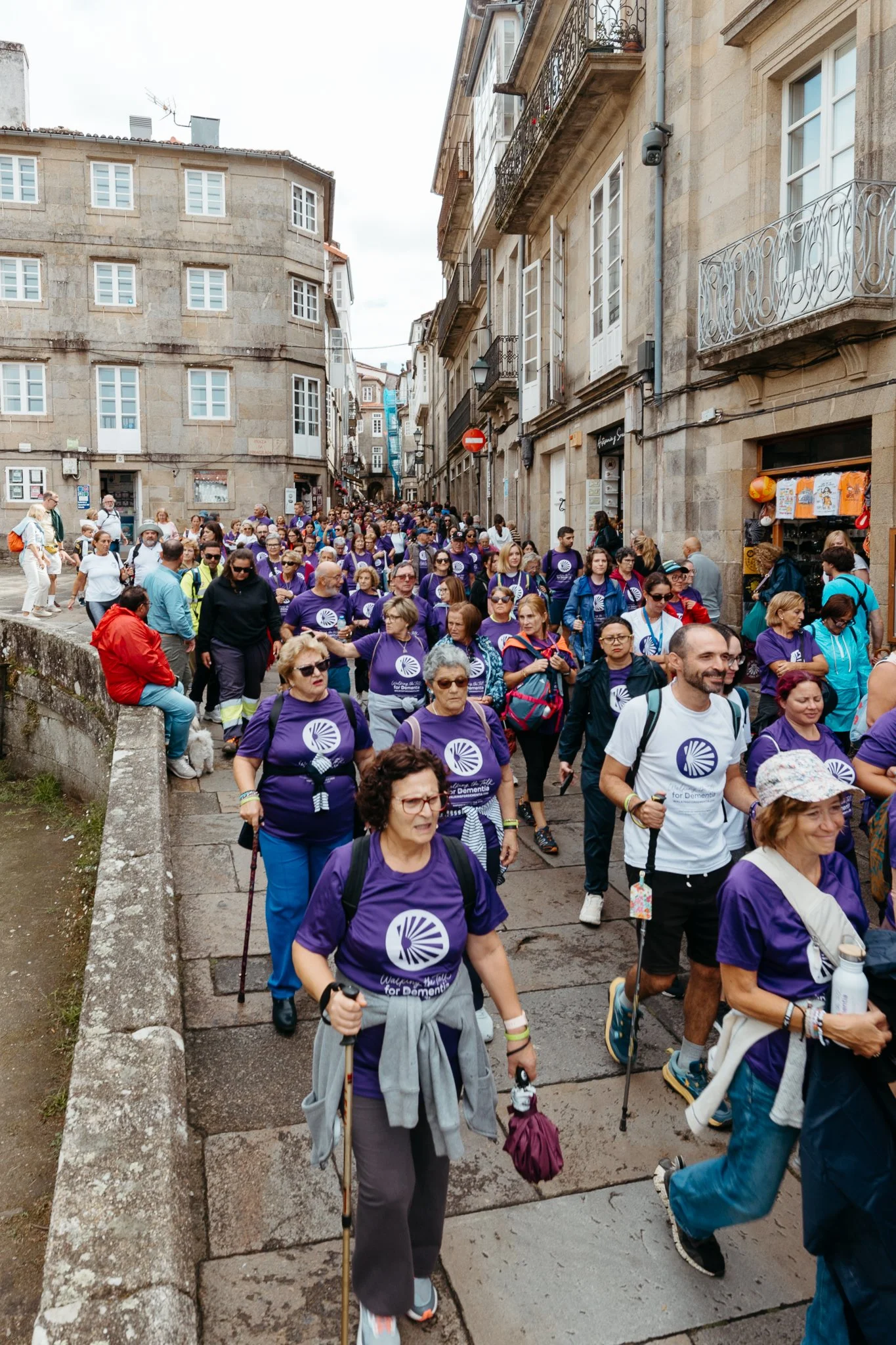 A large group of people participating in an outdoor walk or protest on a city street, many wearing purple shirts with a circular logo, some carrying backpacks and walking sticks, surrounded by old stone buildings with balconies.