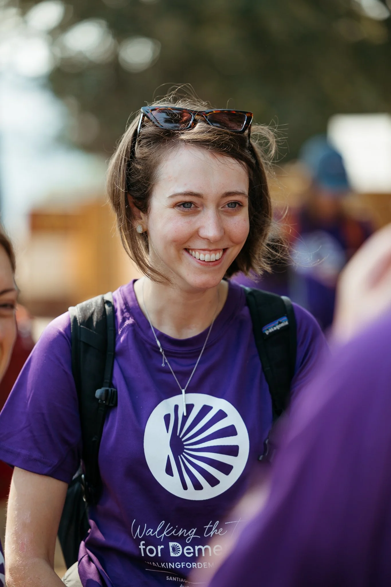 A young woman smiling, wearing a purple t-shirt with the logo and text 'Walking the for women,' sunglasses on her head, and a backpack, outdoors during daytime.