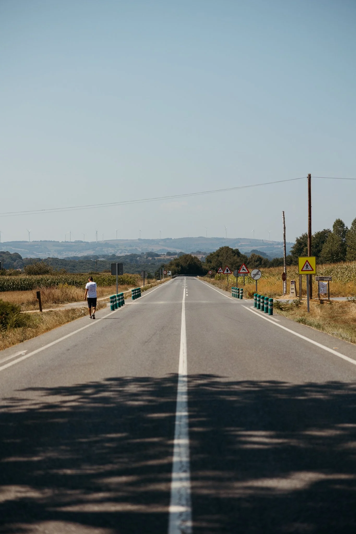 A rural road extending into the distance with a person walking on the left sidewalk and traffic signs along the right side, under a clear blue sky.