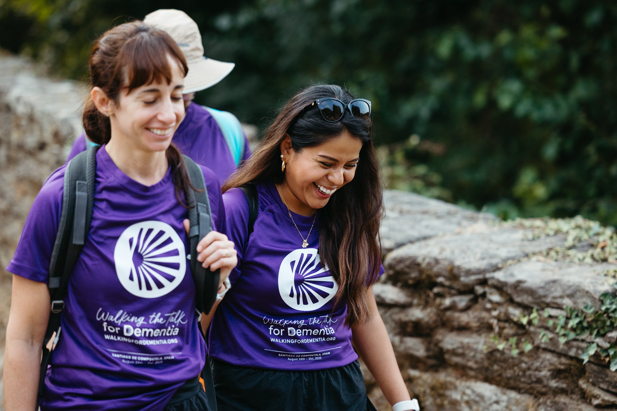 Three women walking outdoors in a forest, wearing purple shirts with a logo and text about walking for dementia.