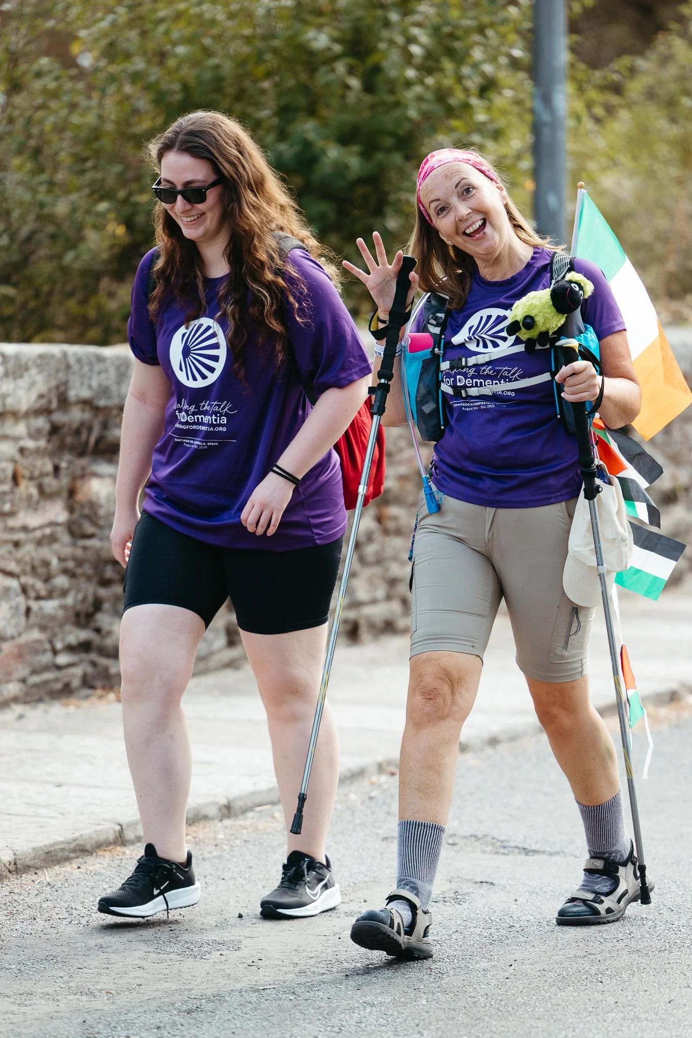 Two women walking outdoors, wearing matching purple shirts with a white logo, carrying backpacks, walking sticks, and smiling. One woman has a rainbow flag attached to her backpack, and the other is wearing sunglasses and smiling.