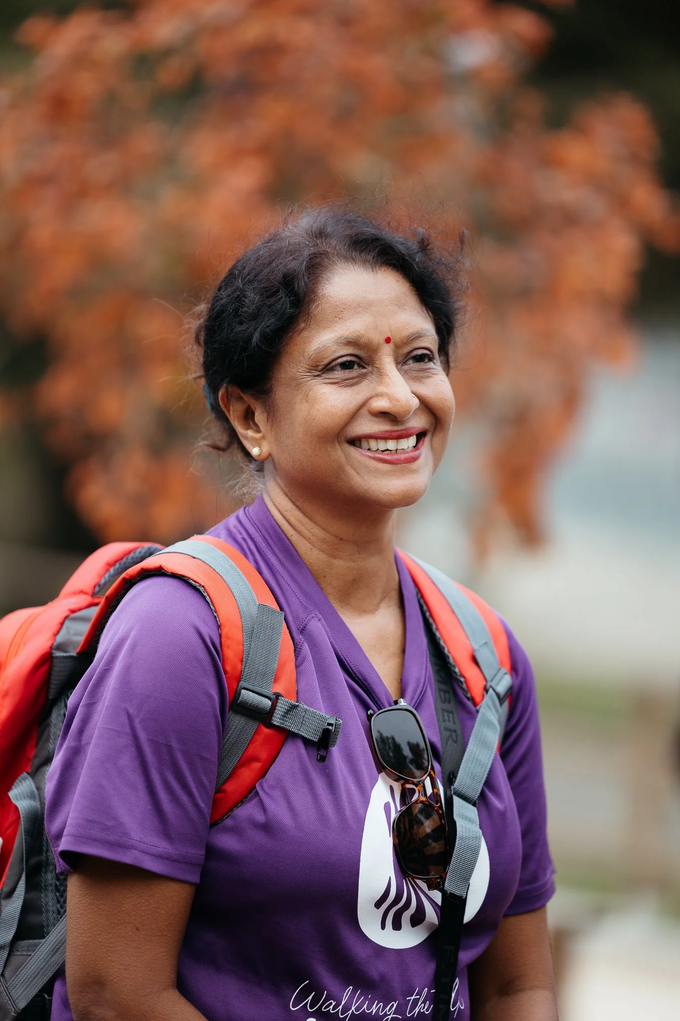 Smiling woman outdoors wearing a purple shirt and a red backpack, with sunglasses hanging from her shirt, against autumn-colored trees.