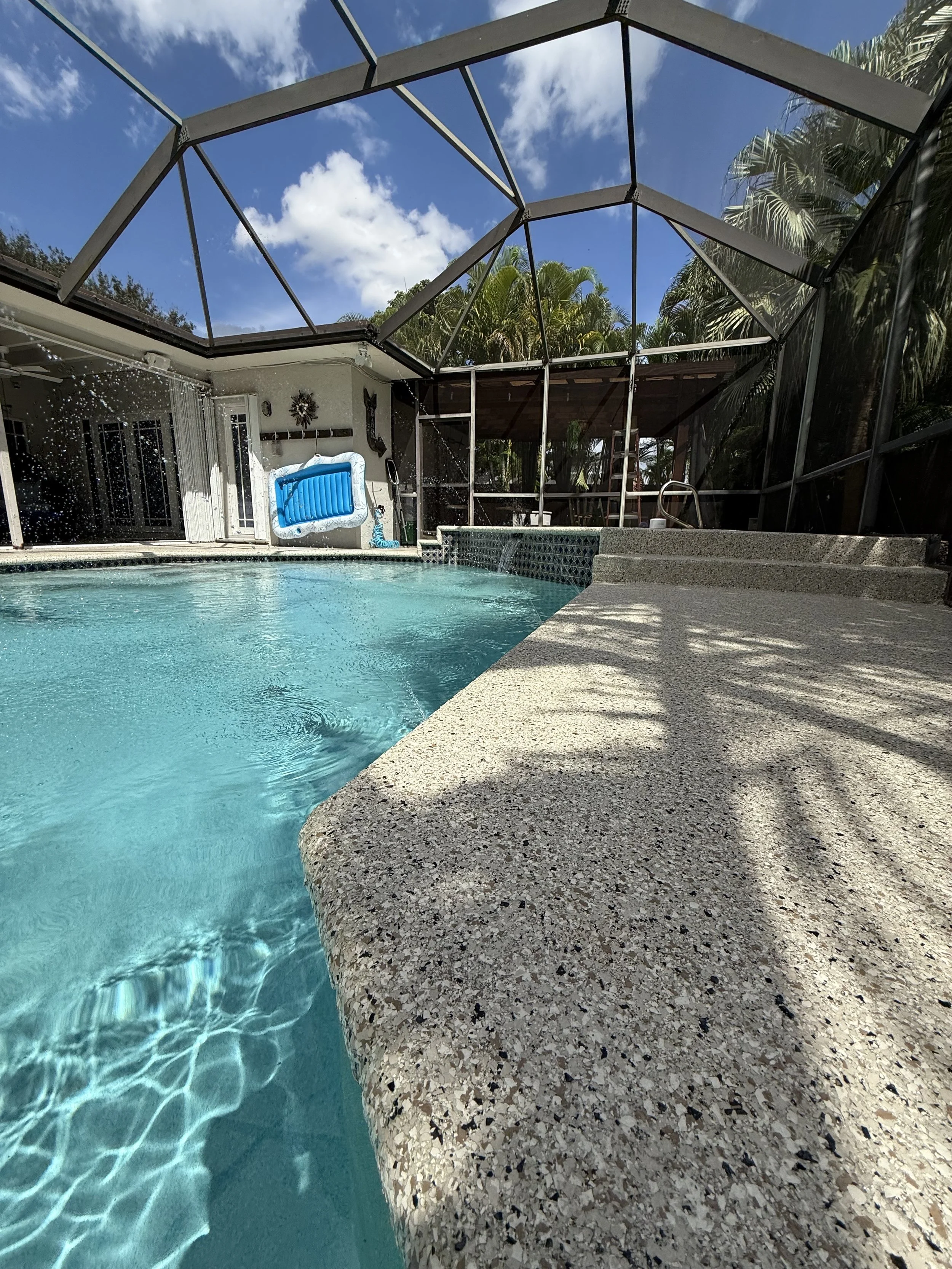 Vista de una piscina en un patio trasero con una estructura de protección de malla metálica, hay sombra en la orilla de la piscina y algunos árboles tropicales y un cielo azul con nubes blancas en el fondo.