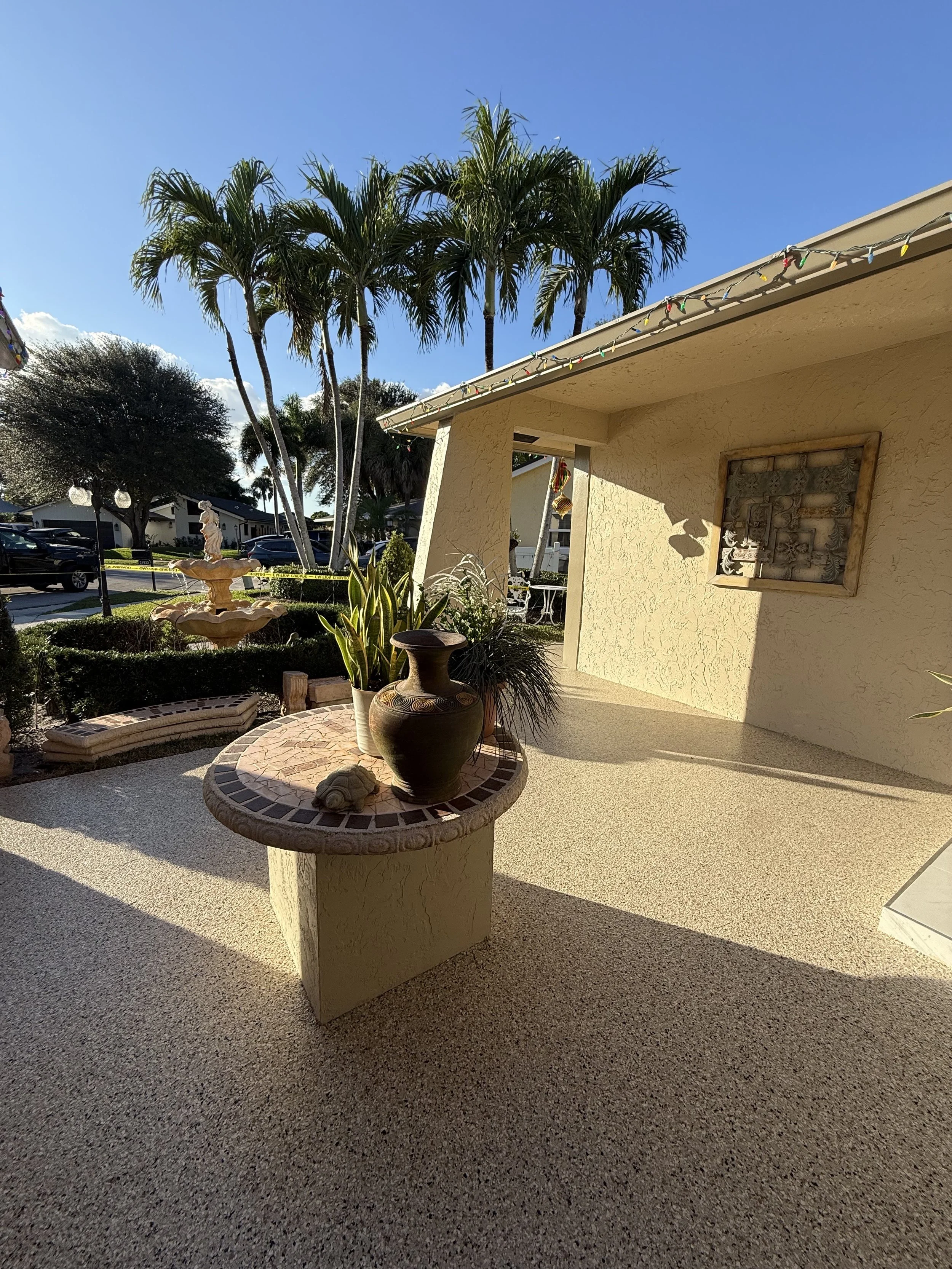A sunny patio with beige stucco walls, a round mosaic-tiled table holding ceramic vases and plants, and palm trees in the background.