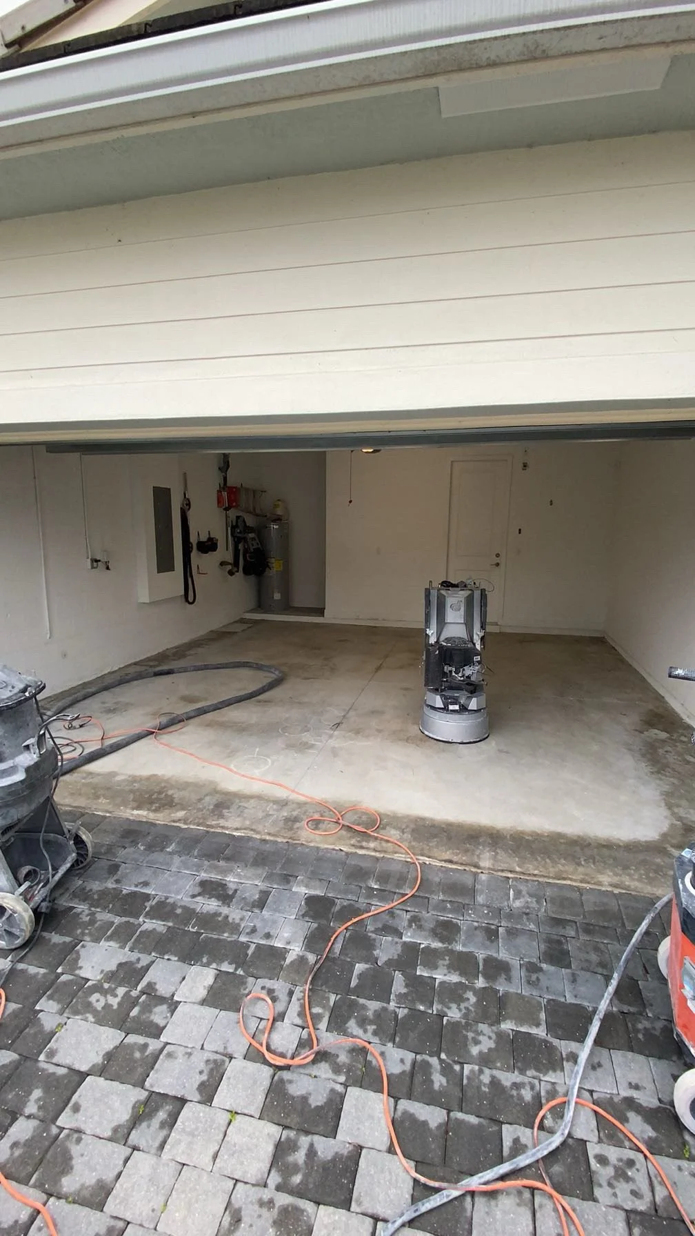 A partially renovated garage with construction tools and equipment. The floor is in the process of being resurfaced, with some areas still showing concrete and others already completed with paver stones. There are electrical cords and a portable vacu