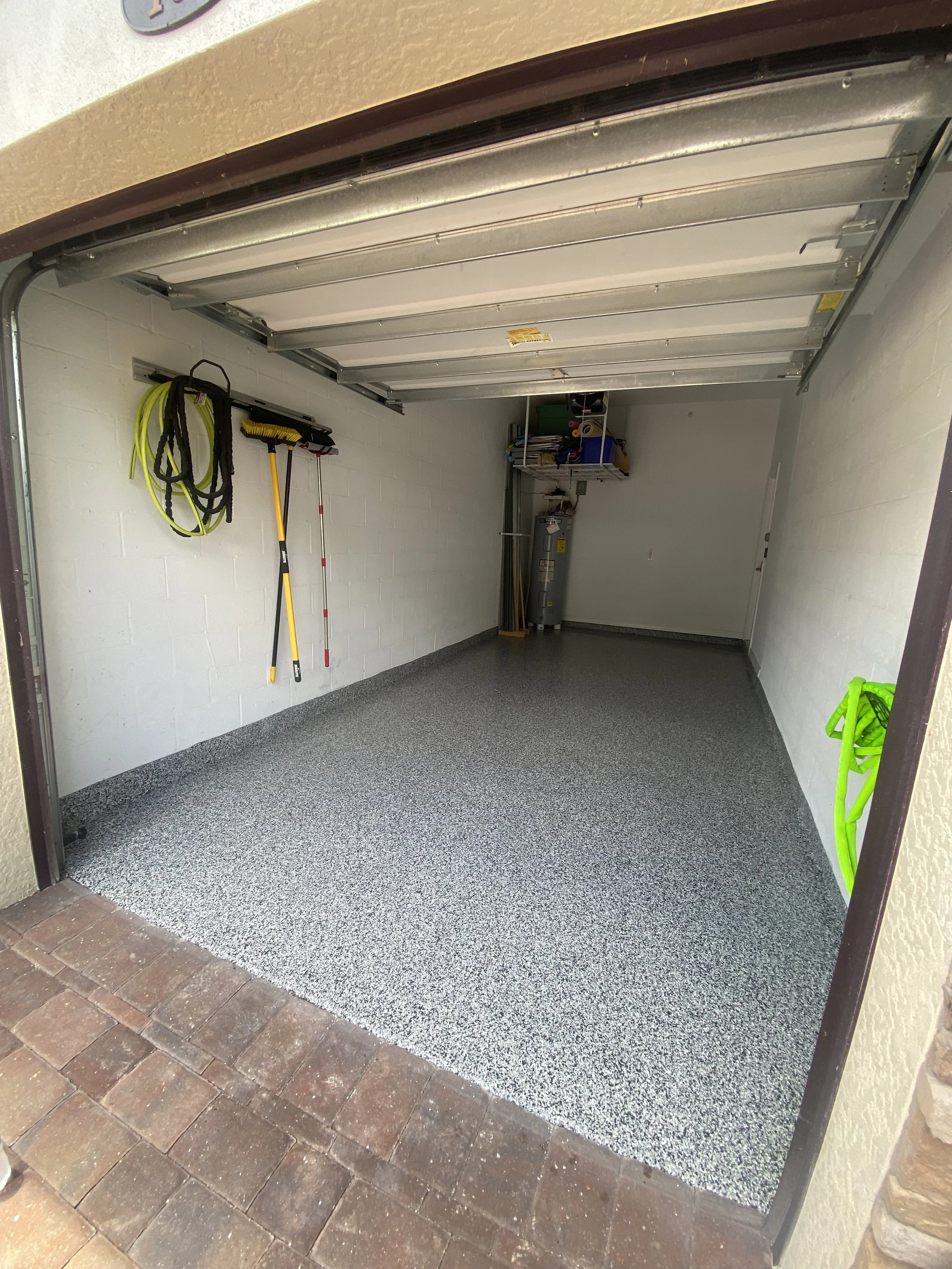 Empty garage with a speckled gray epoxy floor, white painted cinder block walls, and a partially open metal garage door. Tools like a broom, rake, and high-visibility garden hose are hanging on the left wall. The back of the garage has shelves with s