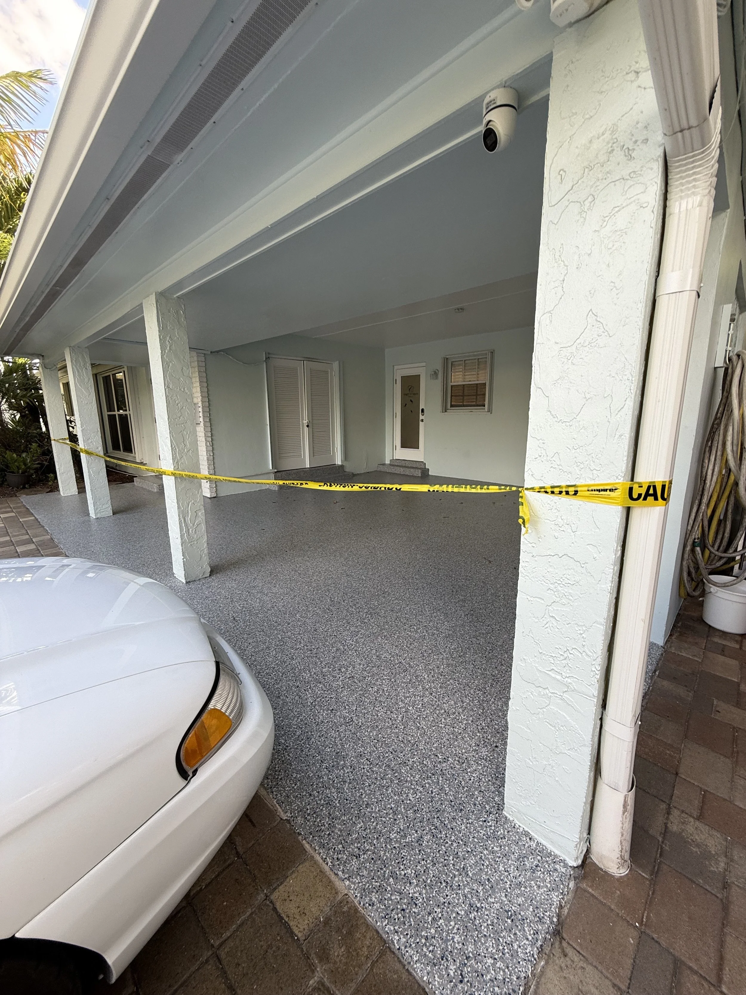 Residential garage with caution tape, a parked white car visible in the foreground, and a light blue house interior with a door and window in the background.