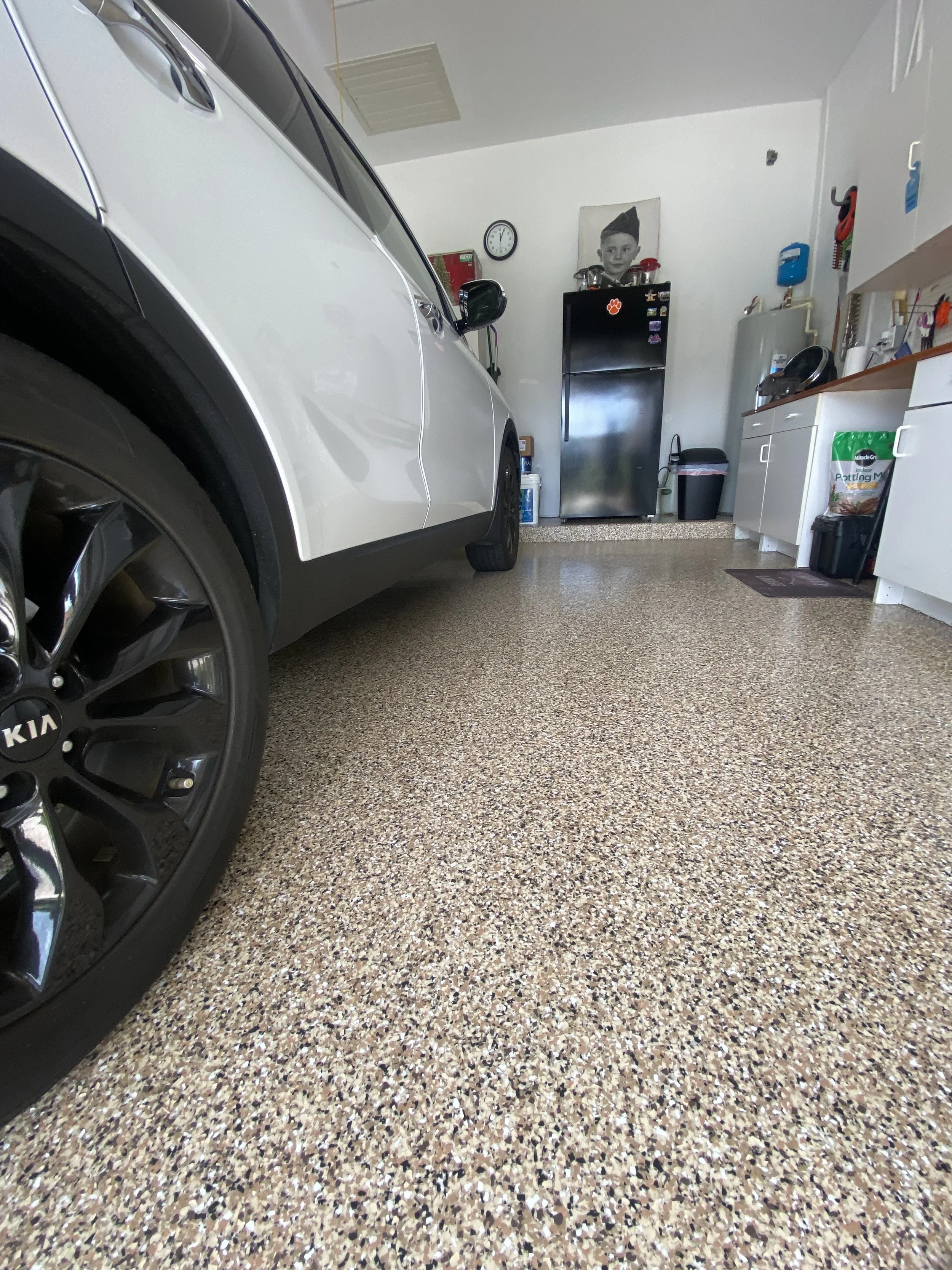 Garage interior with a white Kia vehicle, a black refrigerator, a large black and white portrait on the wall, and various storage cabinets and items.