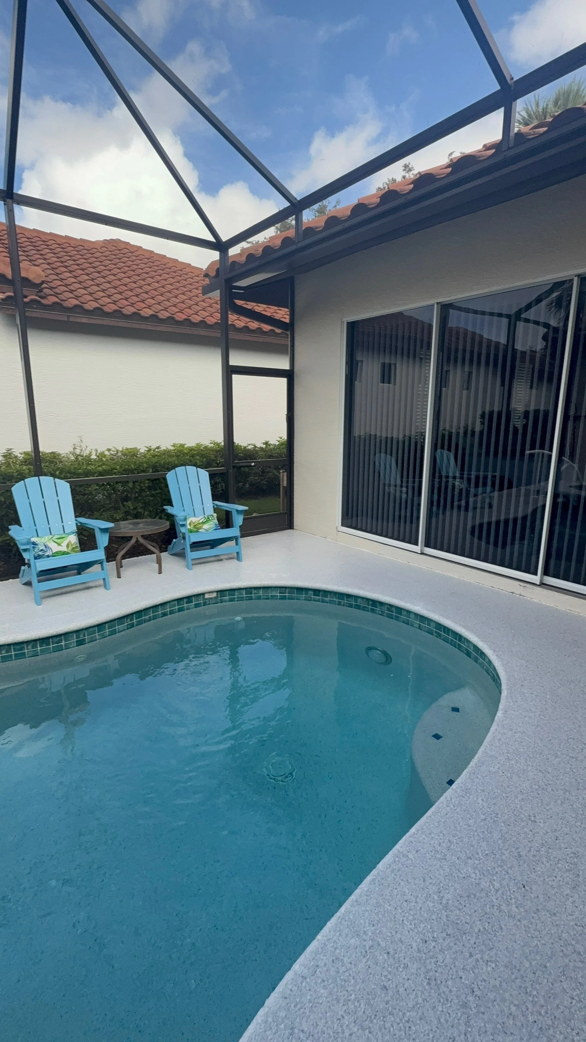 Piscina con vista a una casa con molduras negras, sillas Adirondack azules y cielo parcialmente nublado.
