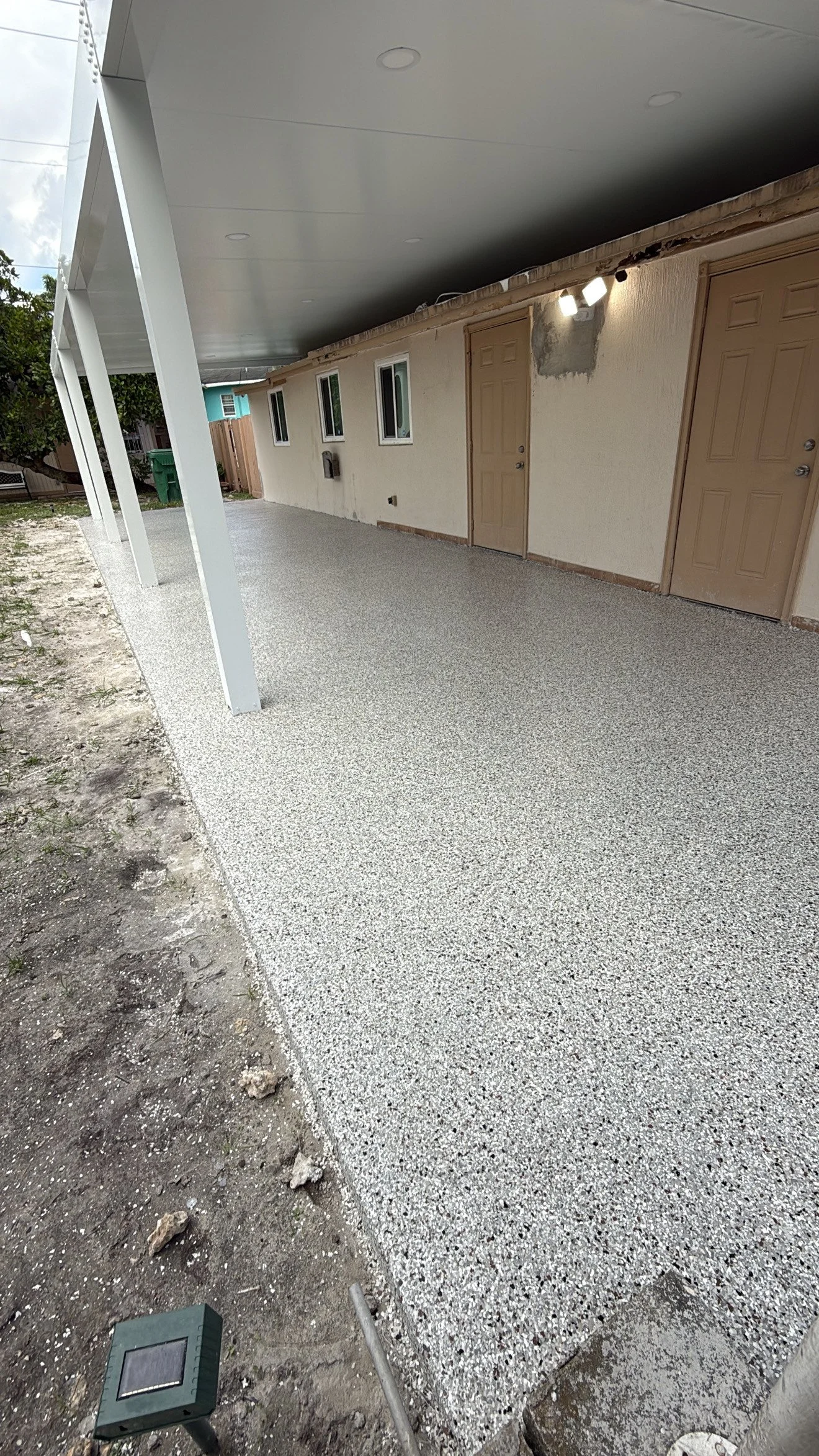 Finished concrete patio with a pebble texture, attached to a house with beige doors and yellow walls, supported by white posts, with a small garden area to the side and utility boxes on the wall.