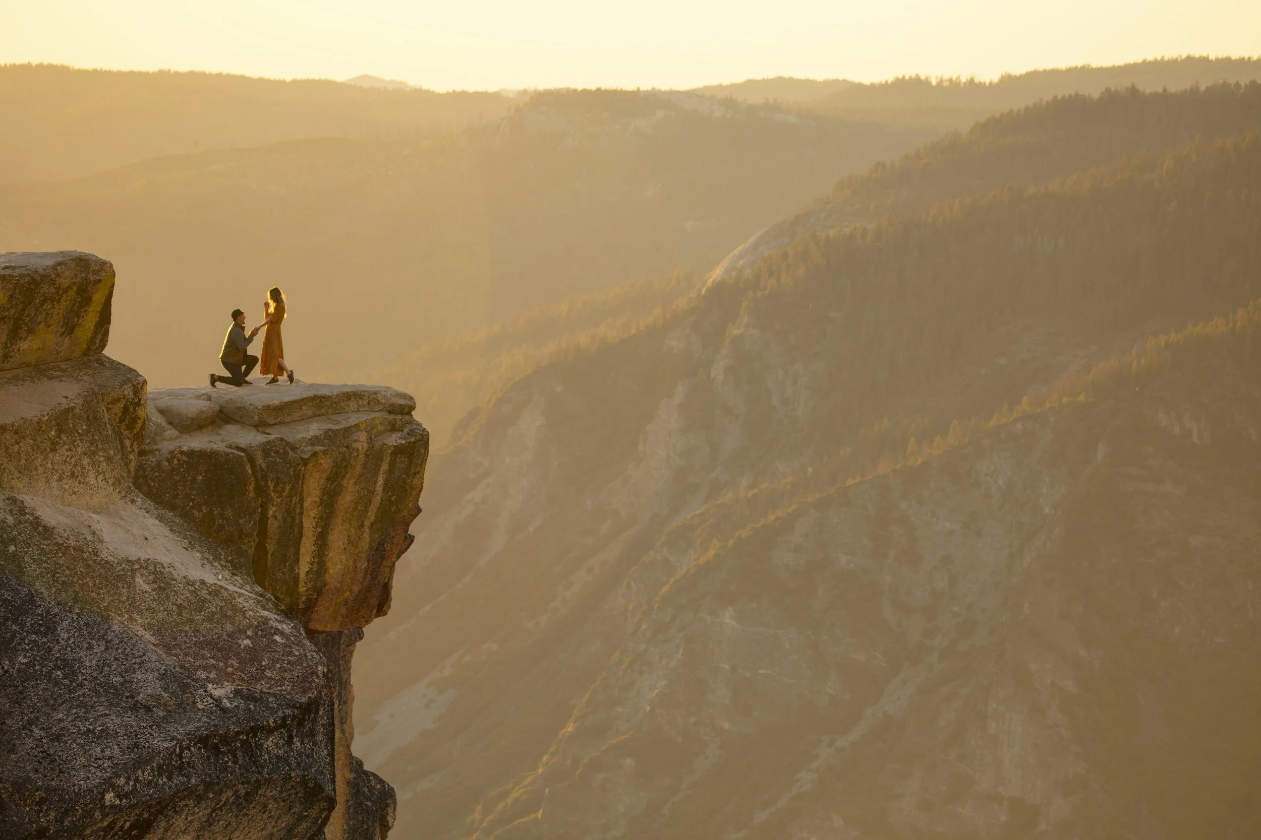 A man proposing marriage to a woman on a rocky cliff at sunset with expansive canyon views in the background.