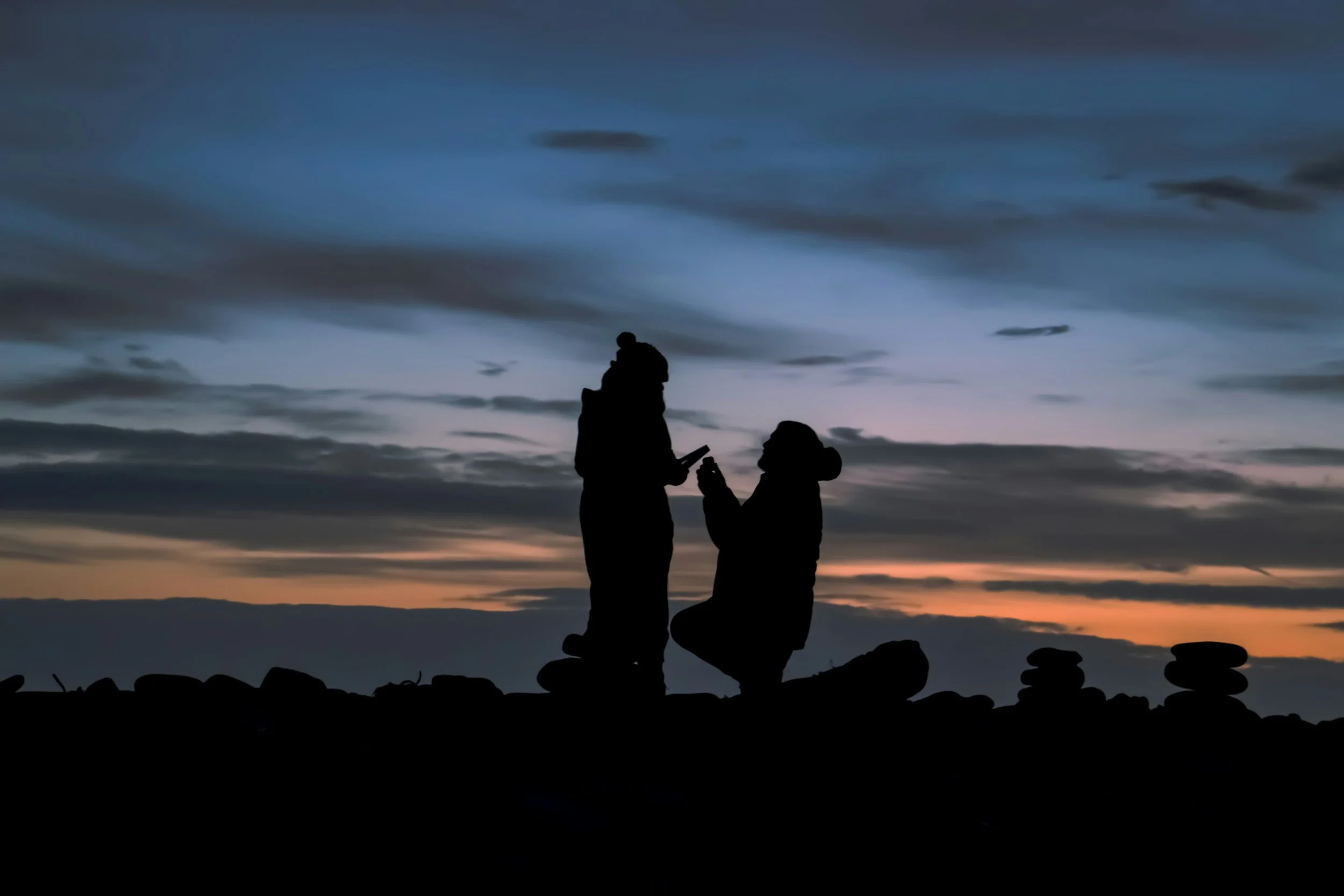 Silhouettes of two people, one kneeling and the other standing, exchanging a small object on a rocky landscape during sunset with a cloudy sky.