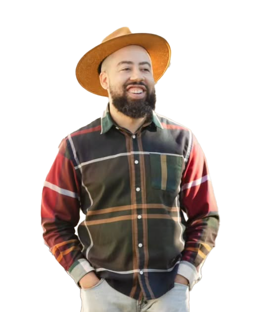 Man wearing a large straw hat and a colorful, checkered button-down shirt, smiling with hands in pockets.