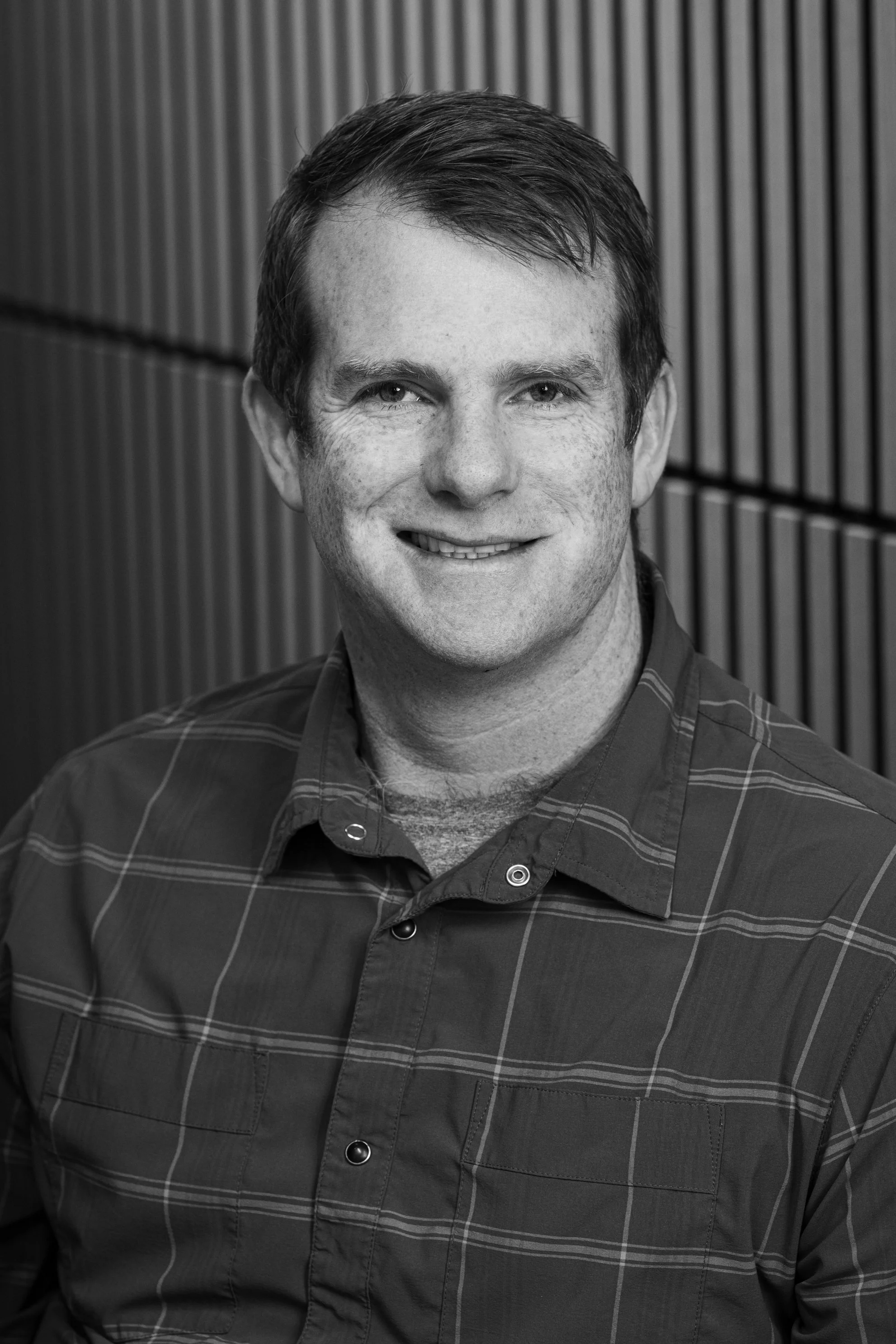 A black and white portrait of a smiling man wearing a plaid shirt, standing against a wooden paneled background.