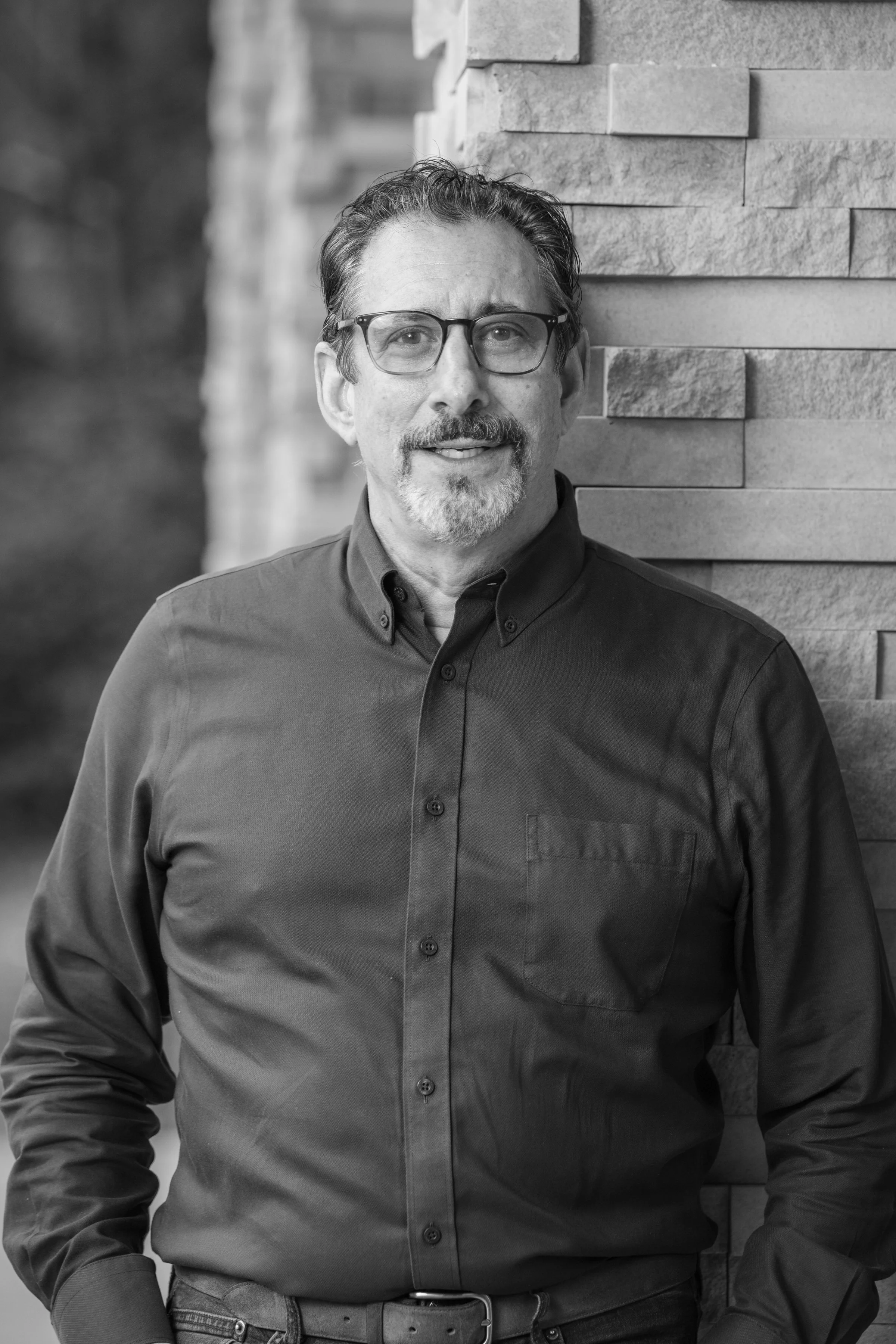 Black and white portrait of a middle-aged man with glasses, a beard, and mustache, wearing a dark button-up shirt, standing against a textured stone wall.