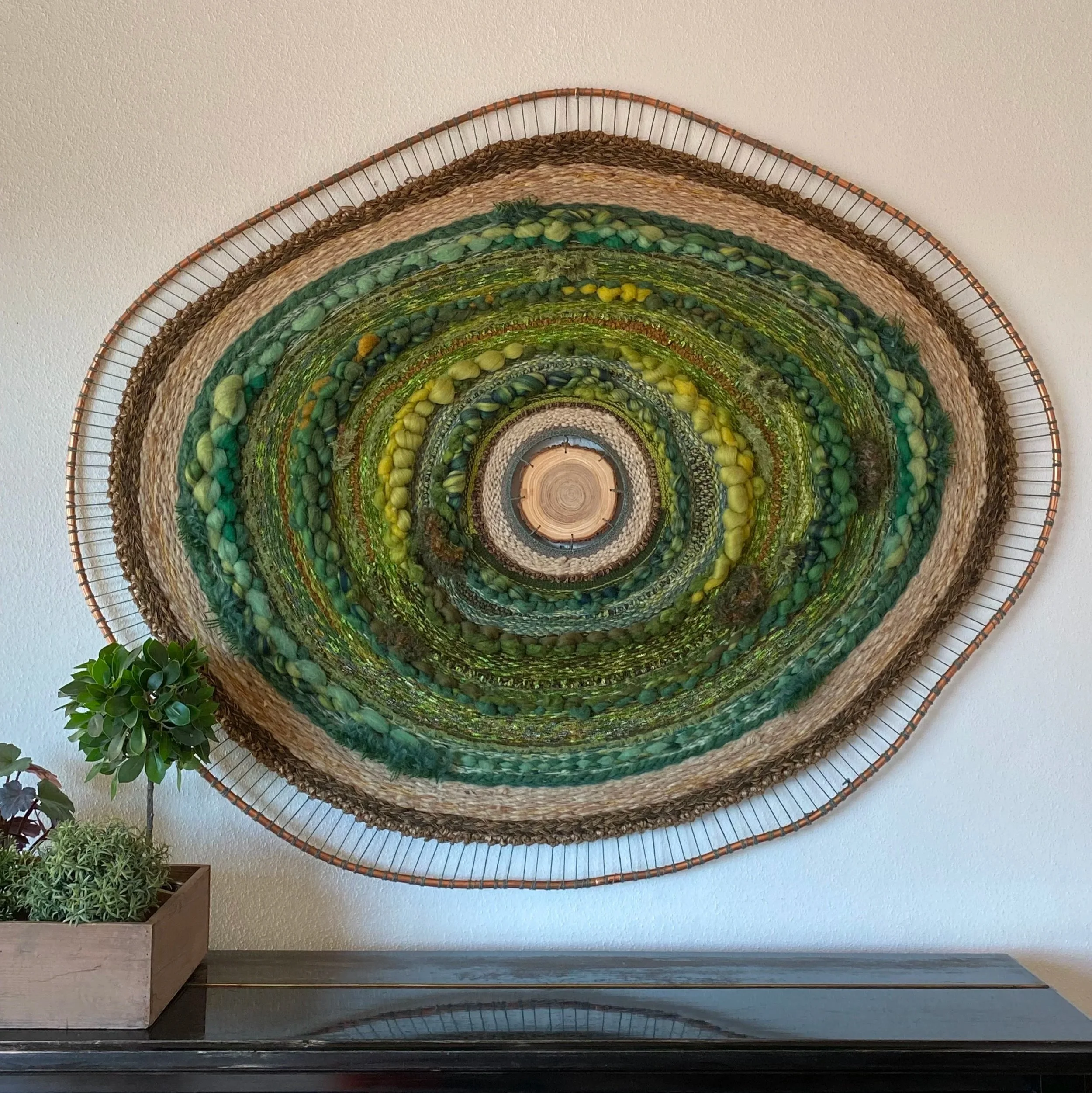 Oval woven wall art with concentric circles in shades of green, brown, and beige, hanging on a white wall above a black surface with a wooden planter containing green plants.