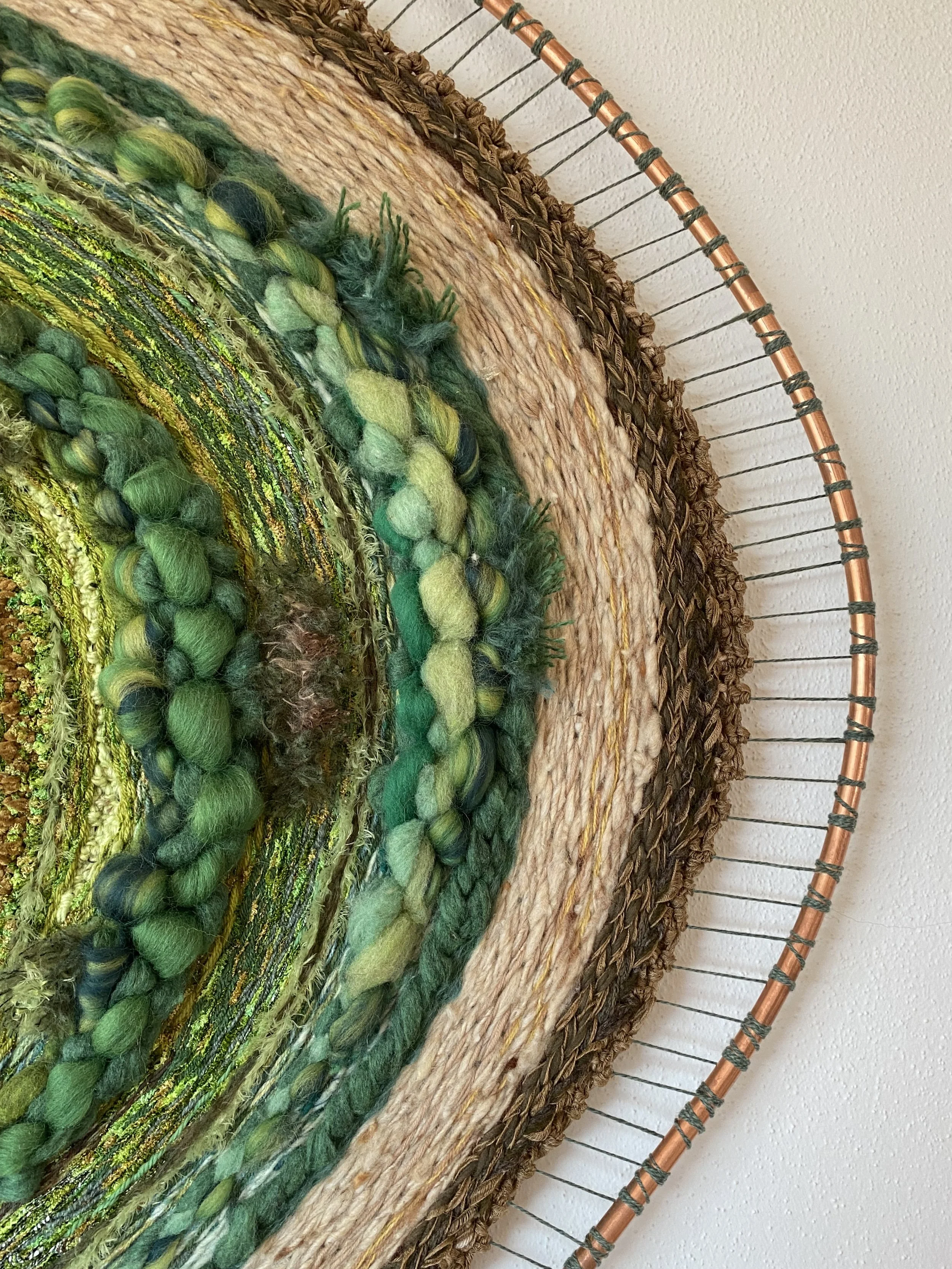 Close-up view of a textured, circular wall hanging made of various woven, knotted, and braided green, yellow, brown, and beige fibers and yarns, with a wire frame around the edge.