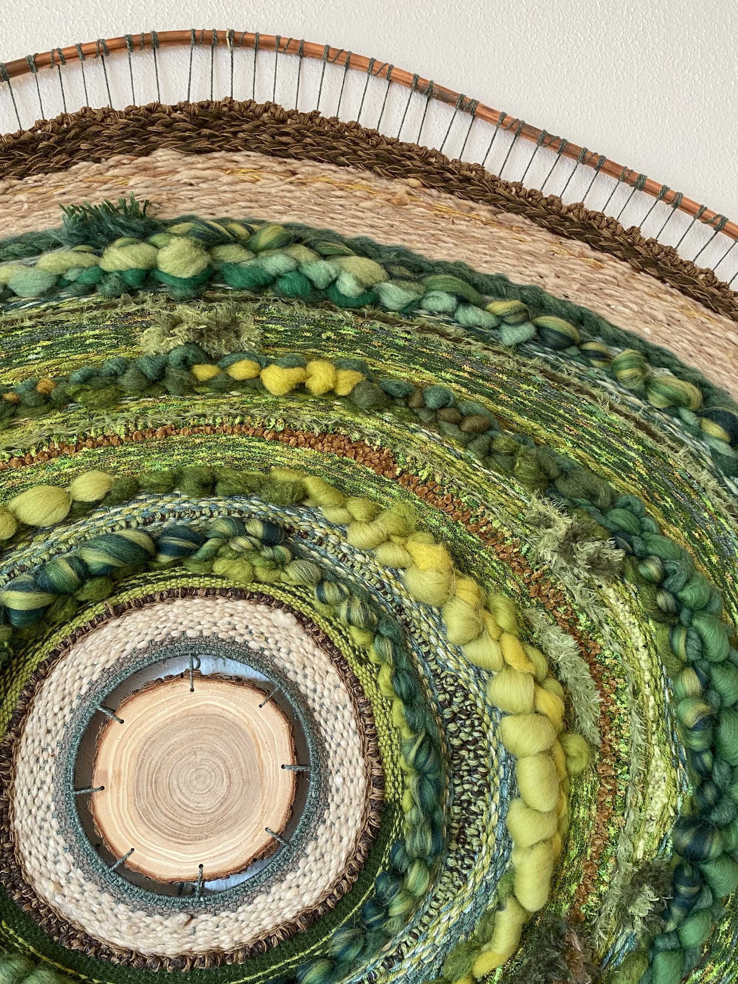 A colorful, layered circular wall hanging made of various felted wool, yarn, and woven materials, with a wooden center at the bottom.