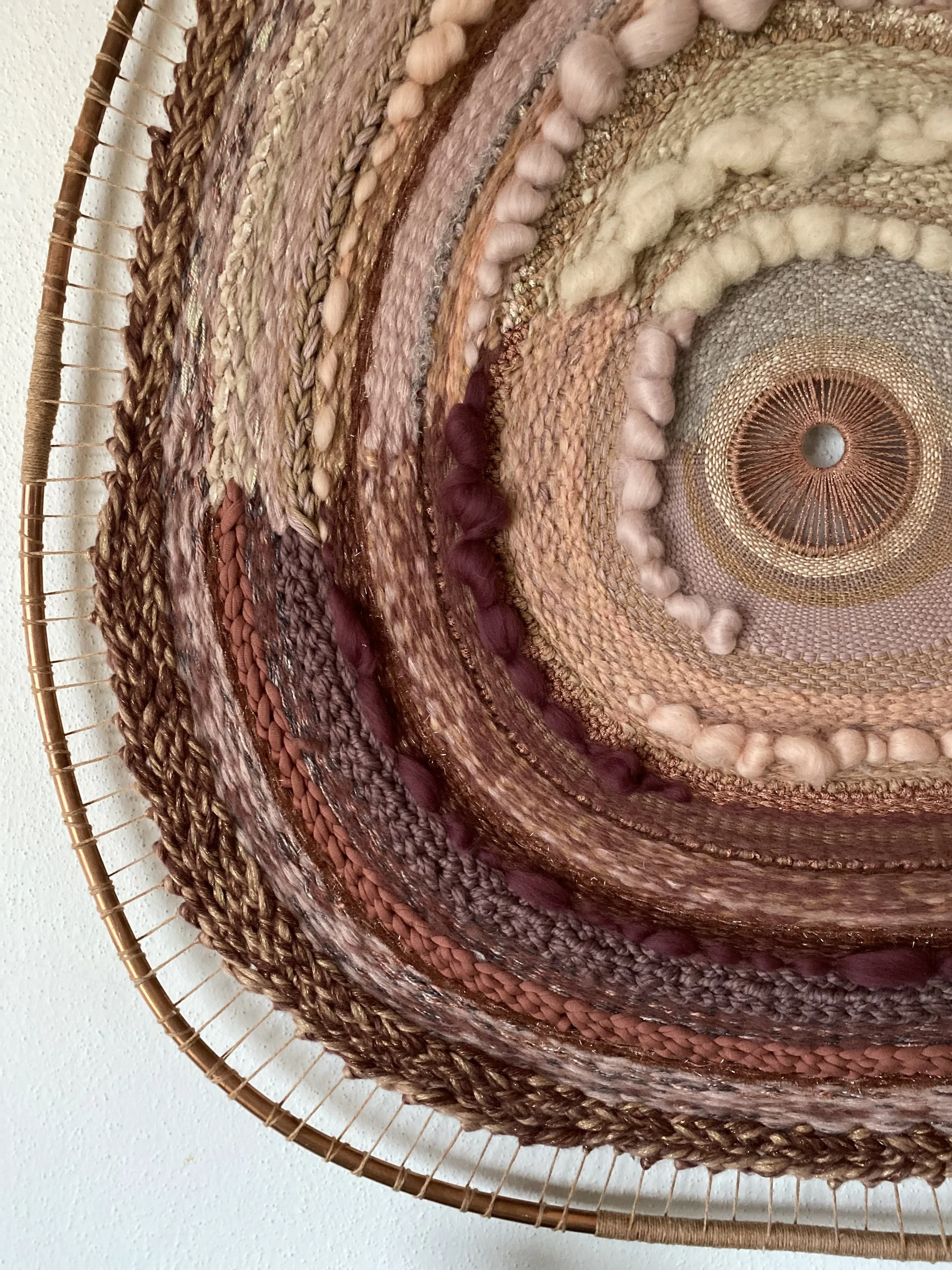 Close-up of a circular woven wall art featuring various textures and colors in shades of brown, beige, pink, and maroon.
